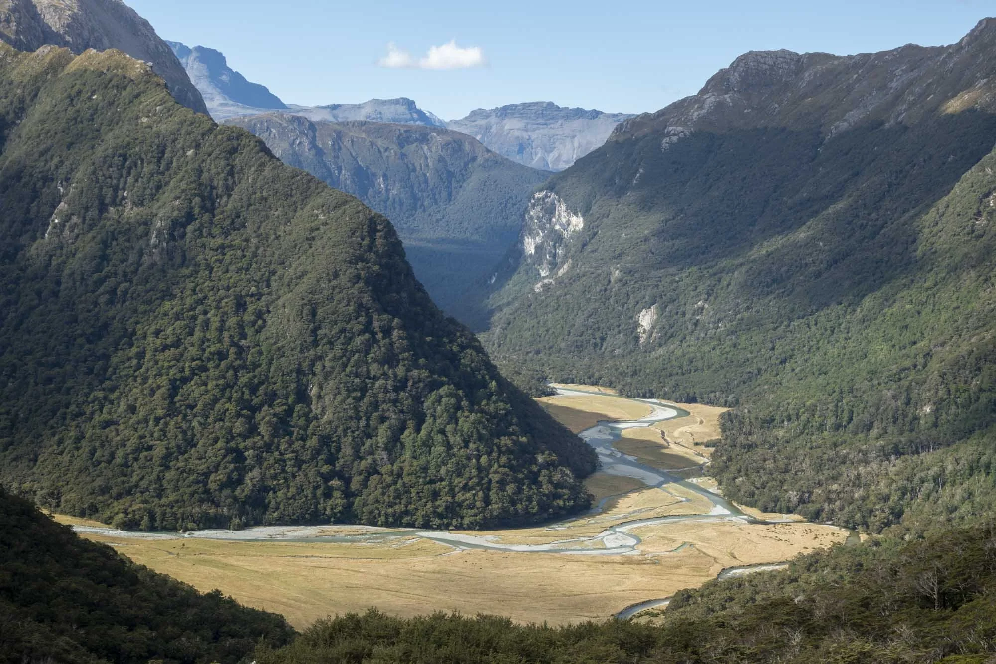 Scenic view of a river winding through a valley with tall, green mountains on each side under a blue sky with scattered clouds.