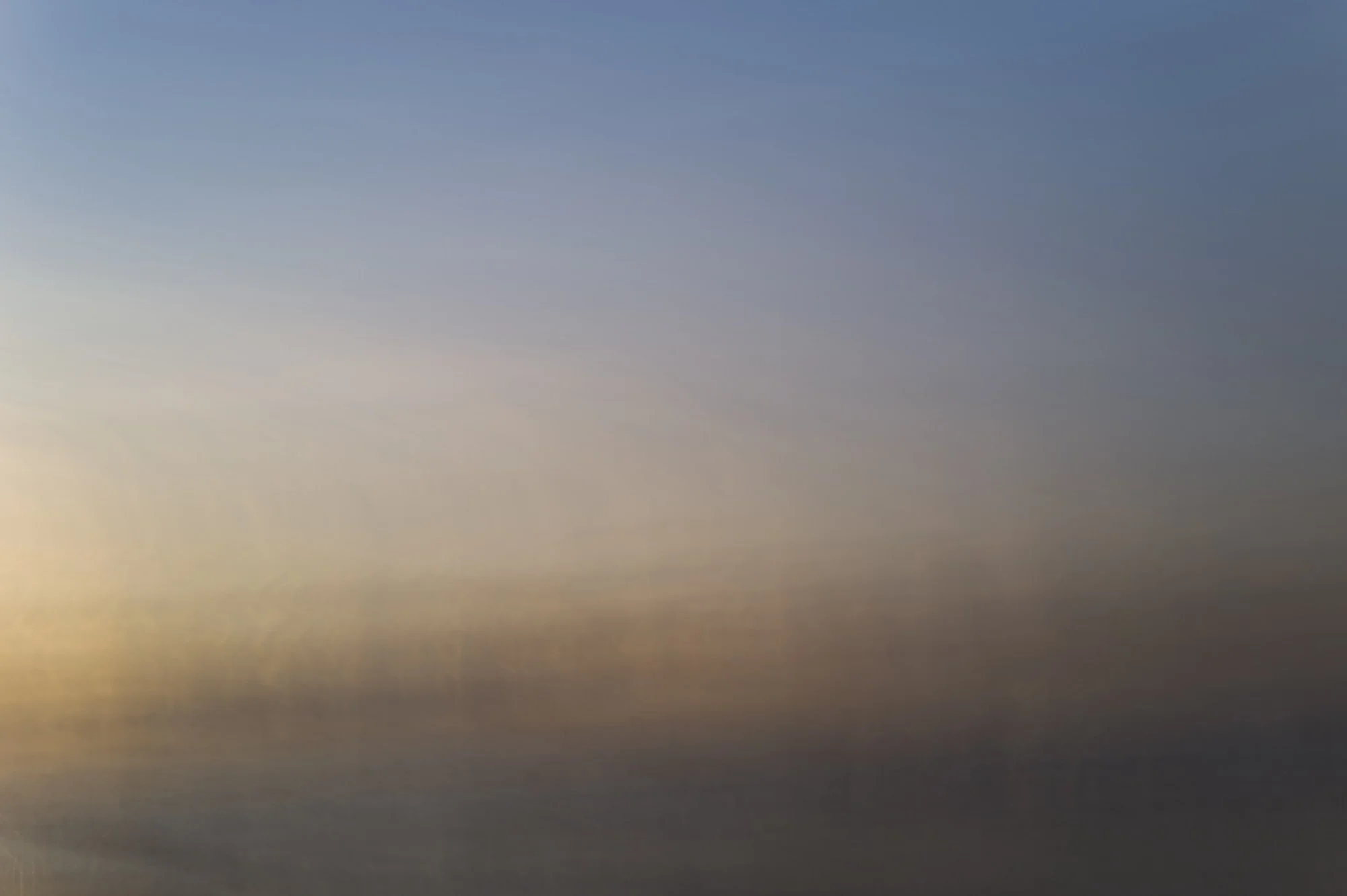 A blurred photograph of a sky during sunset or sunrise with soft pastel colors.