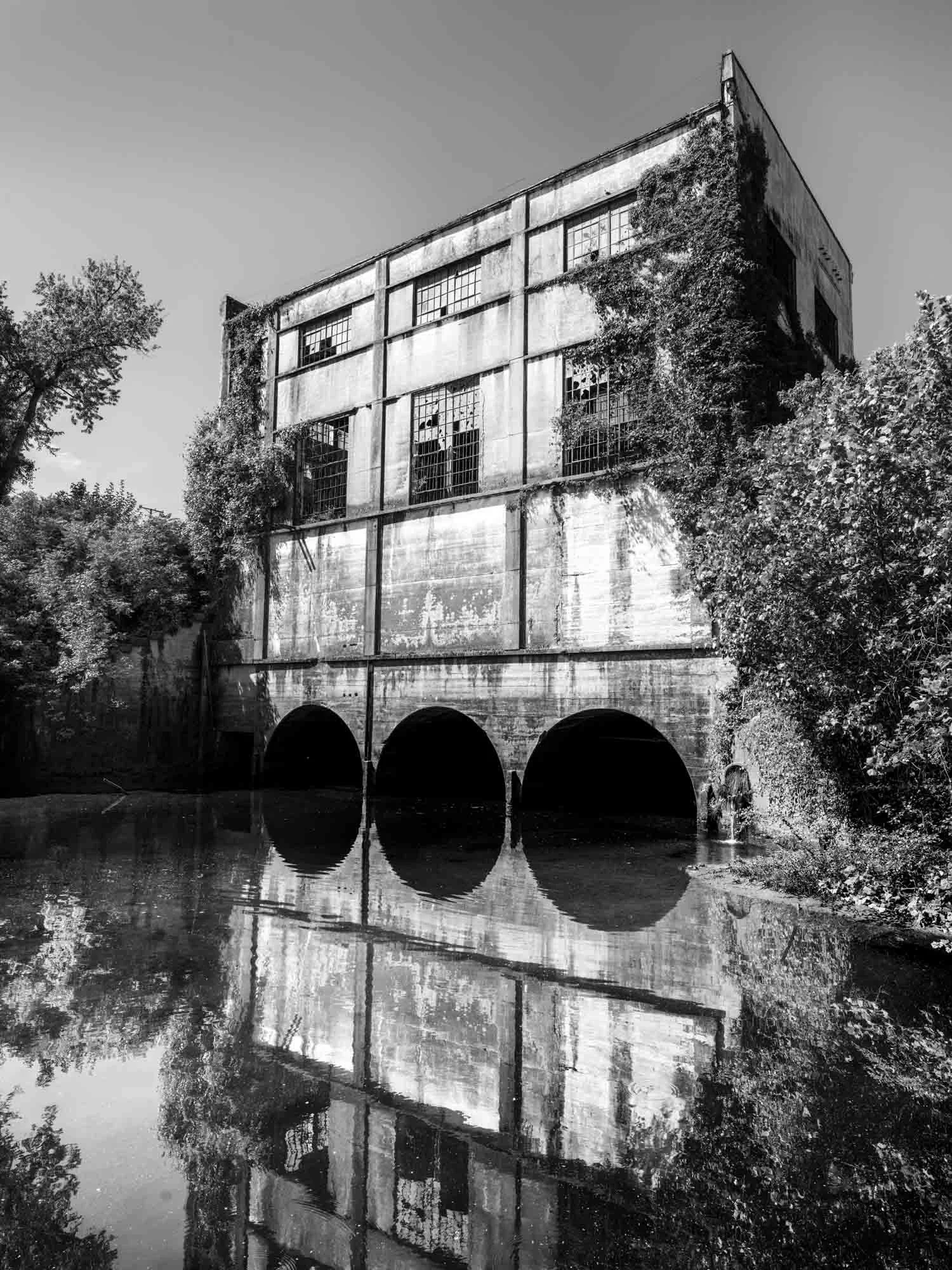Black and white photo of an abandoned industrial building with broken windows, surrounded by trees, reflected in the water below.