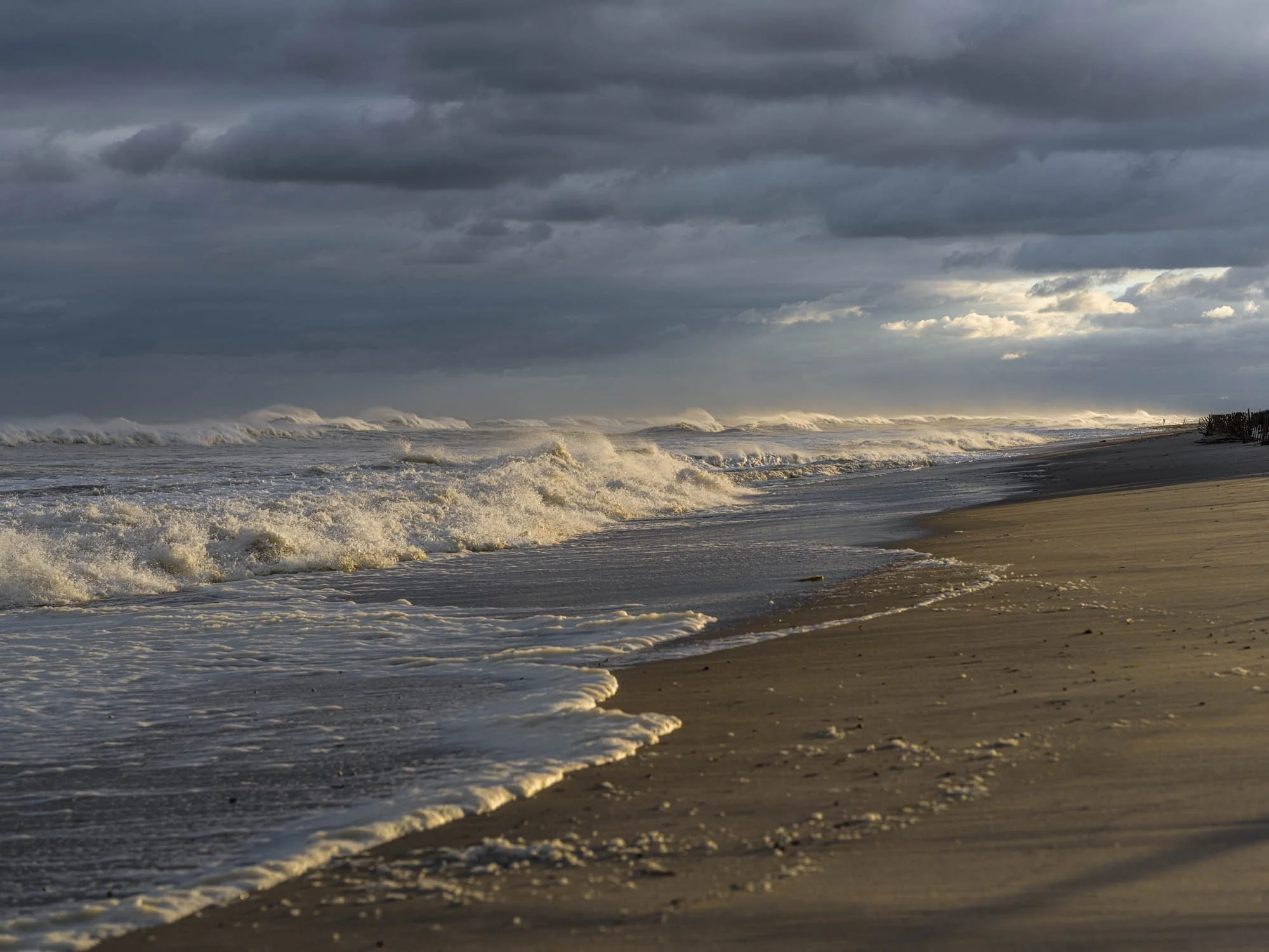Cloudy beach scene with rough waves crashing onto sandy shore and a fence on the right in the distance.