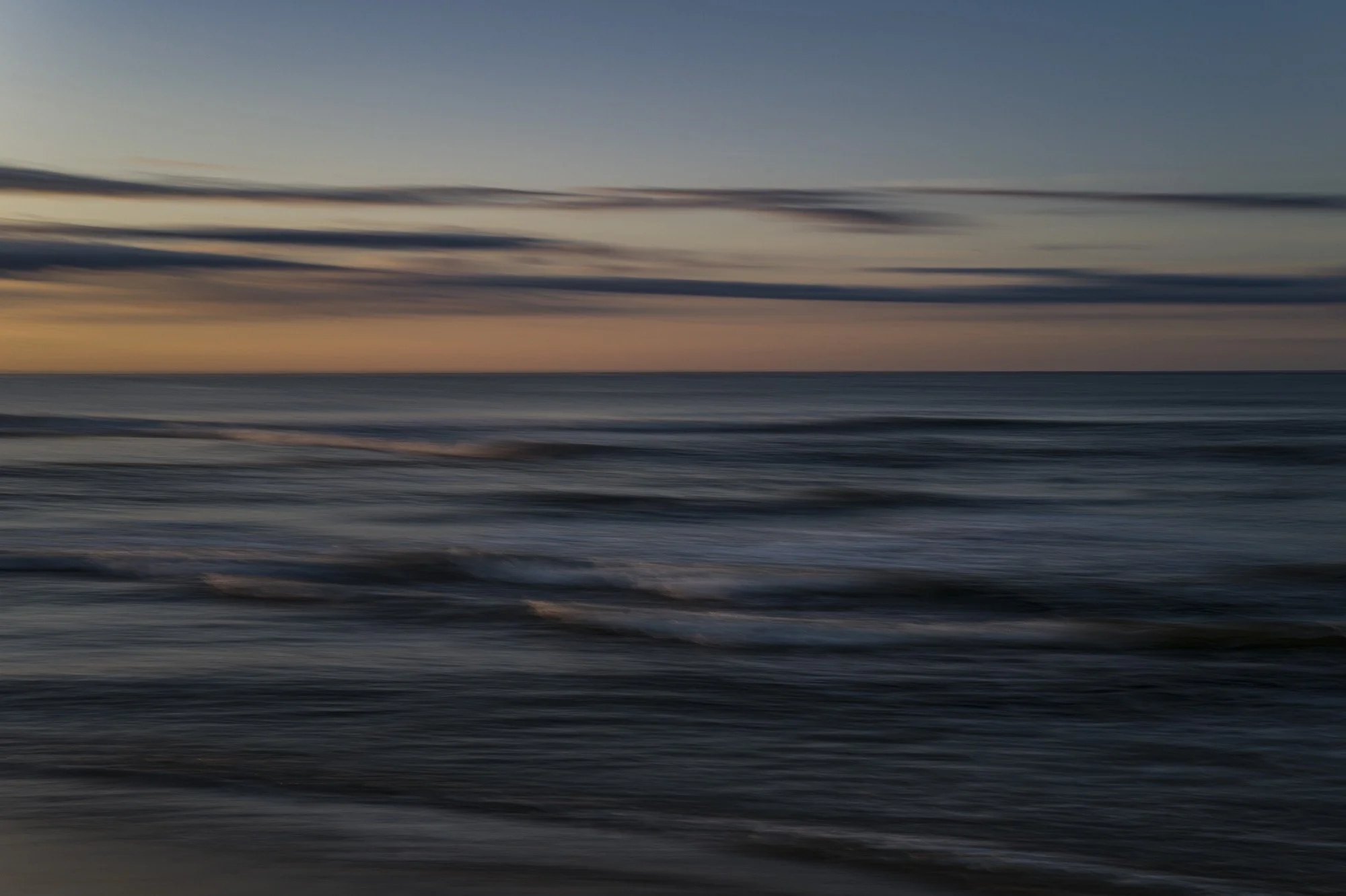 Blurry image of ocean waves at sunset or sunrise with clouds in the sky.