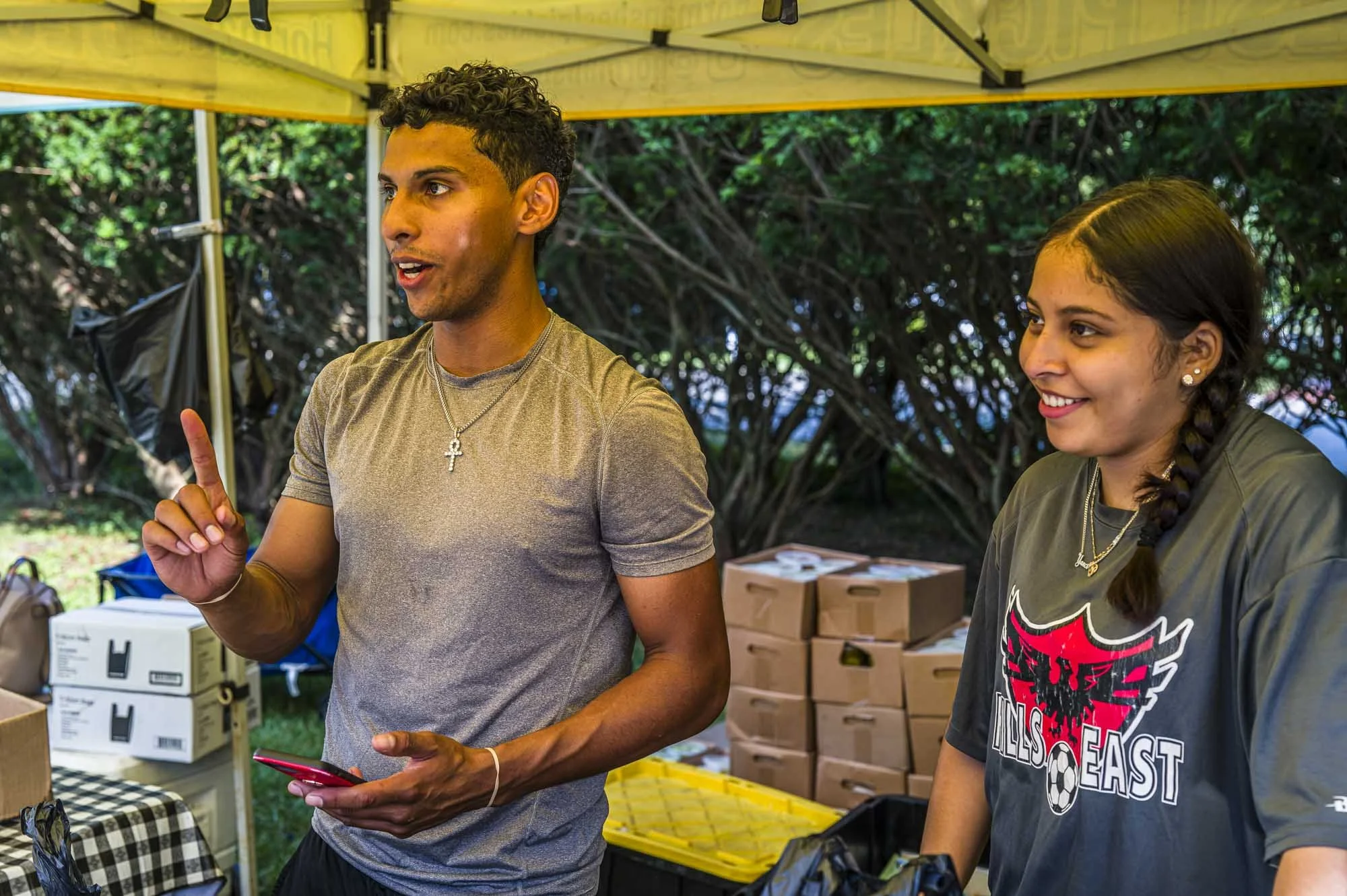 Two young adults, a man with short curly hair and a woman with long braided hair, are standing under a yellow canopy, engaged in conversation. The man is gesturing with his index finger, holding a smartphone in his other hand, and wearing a gray t-sh
