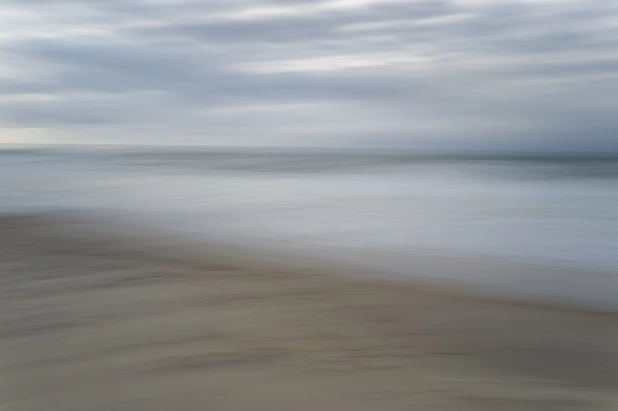 Blurred image of a beach scene with sand, ocean waves, and a cloudy sky.