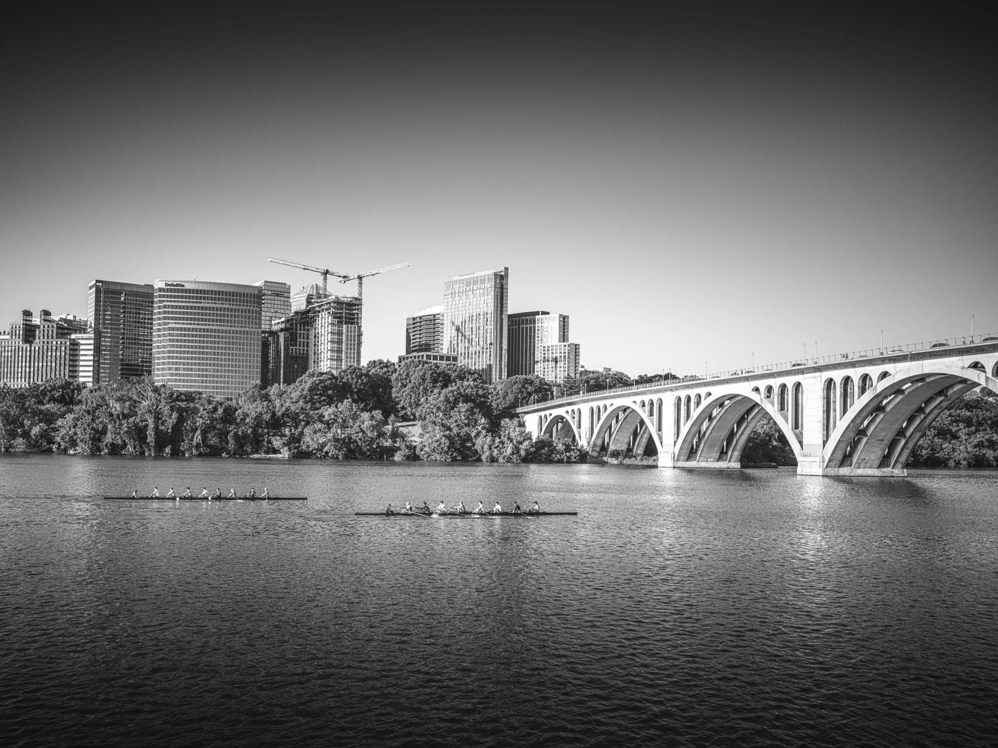 Black and white photo of a city skyline with tall buildings, a large arched bridge over a body of water, and two rowing teams passing on the water.