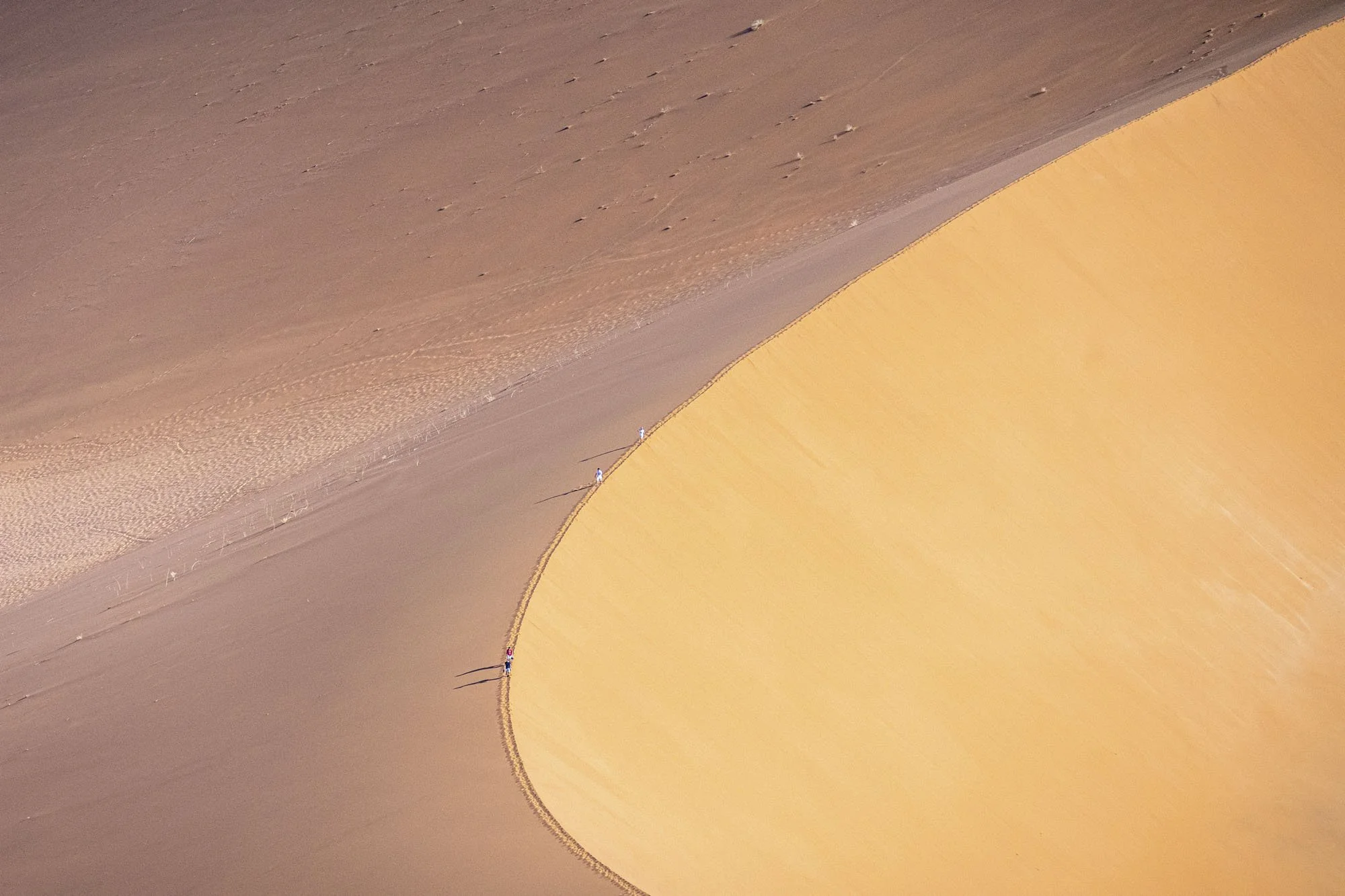 Two hikers walking along a trail beside a massive sand dune in a desert landscape, with long shadows cast by the sunlight.