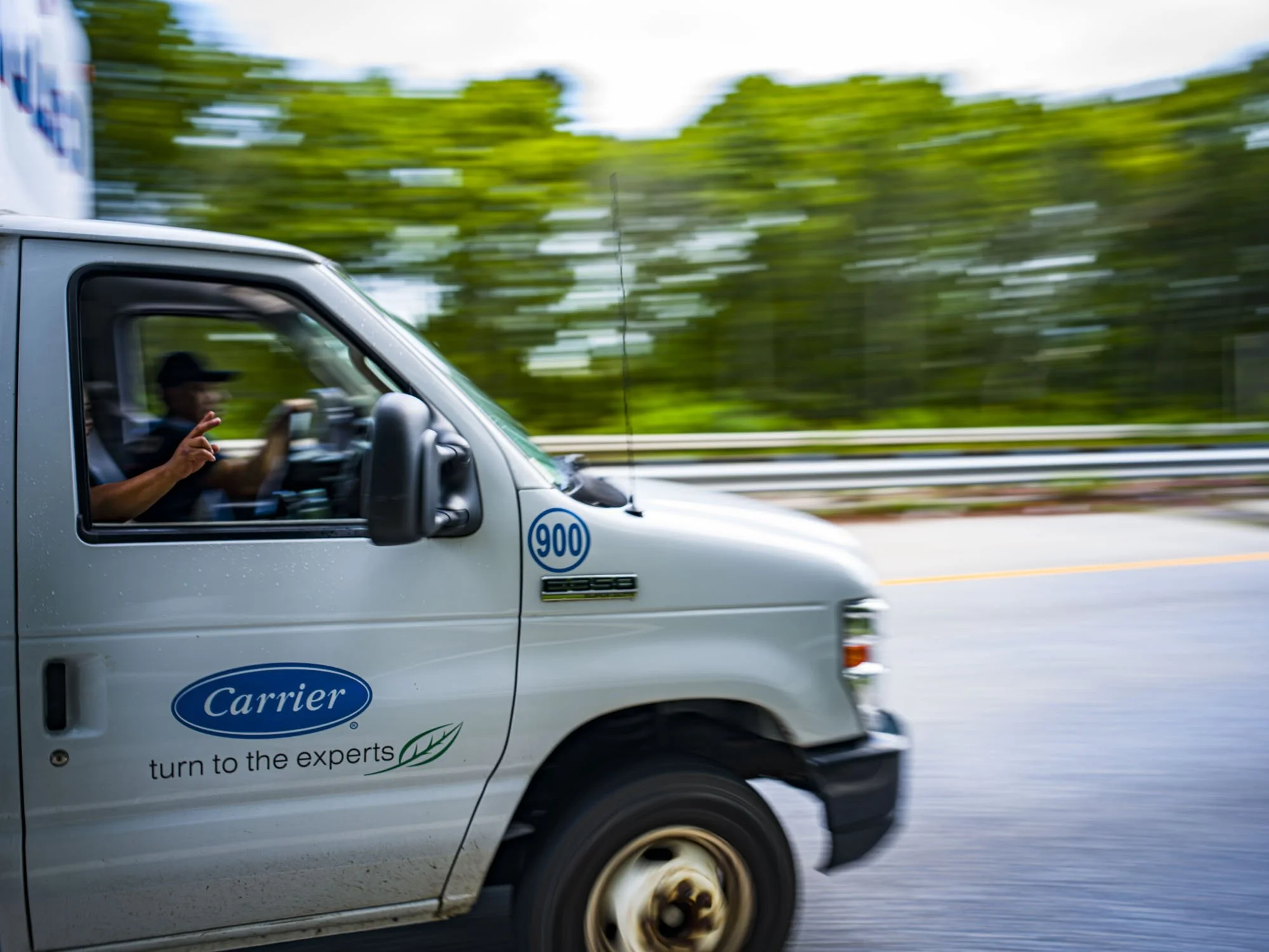 Moving white delivery van with Carrier logo and slogan on side, driver visible through window, background blurred indicating motion.