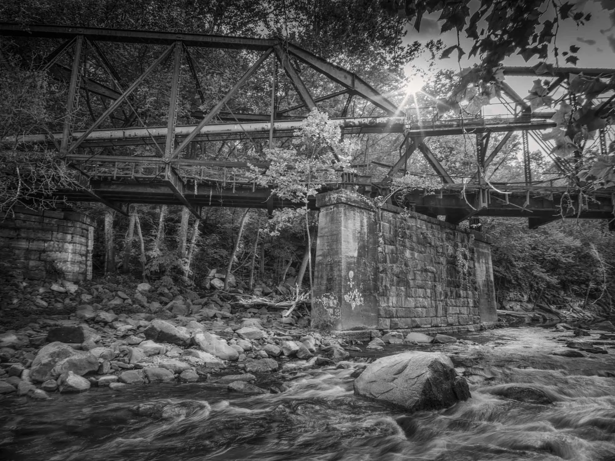Black and white photo of an old, rusted bridge over a rocky stream in a wooded area, with the sun shining through the trees.