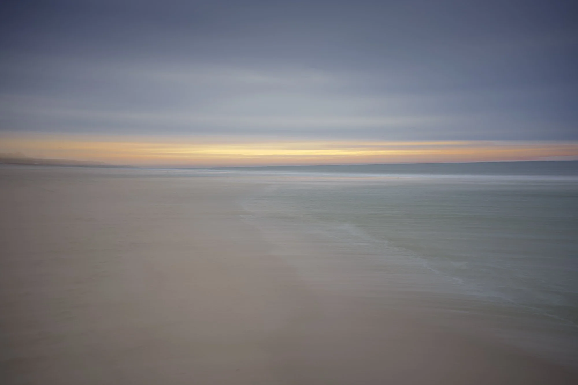 Blurry image of a beach with ocean waves and a colorful sunset or sunrise sky.