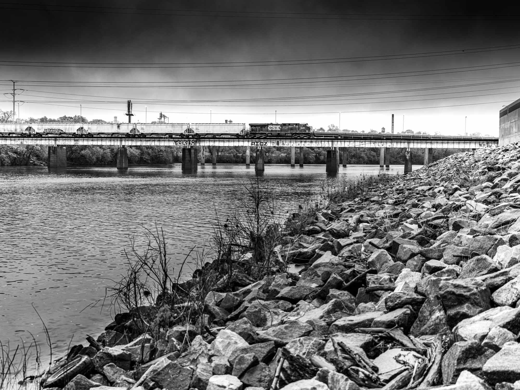 A black and white photograph of a riverbank with a rocky shoreline in the foreground. A train is crossing a bridge over the river, and power lines run above the scene. The sky appears dark and cloudy.