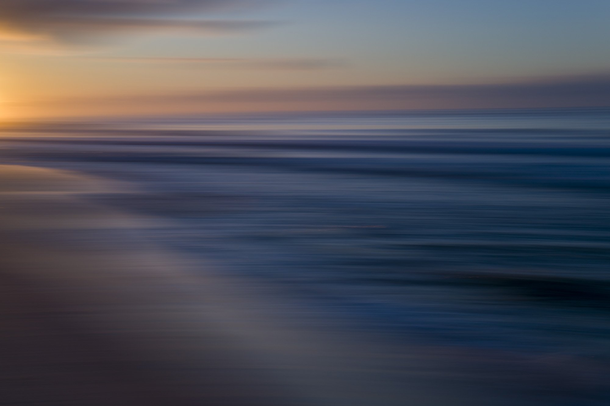 Blurred image of a beach at sunset, showing the horizon, ocean waves, and colorful sky.