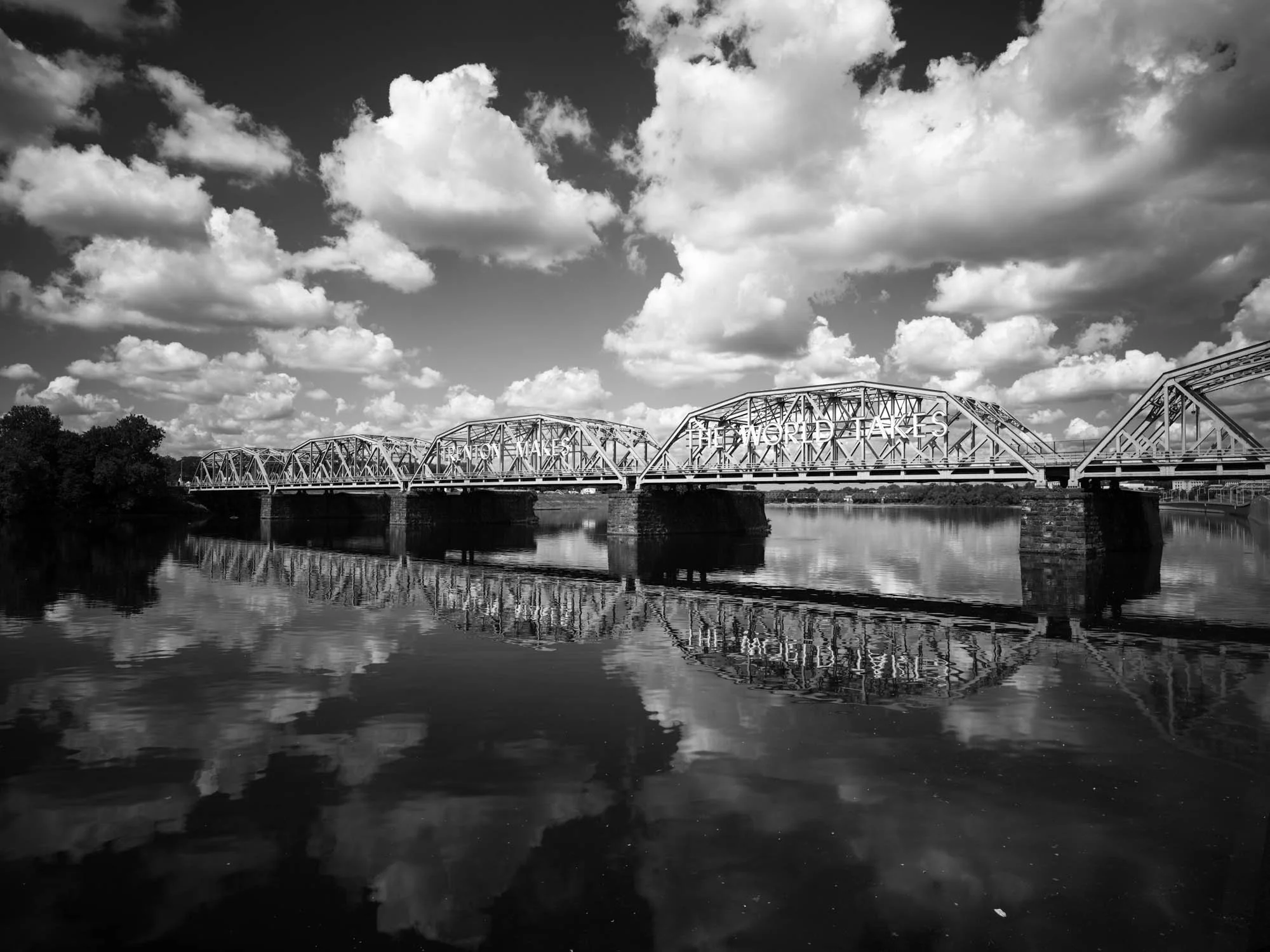 Black and white photo of a bridge over a river with clouds in the sky and their reflection in the water.