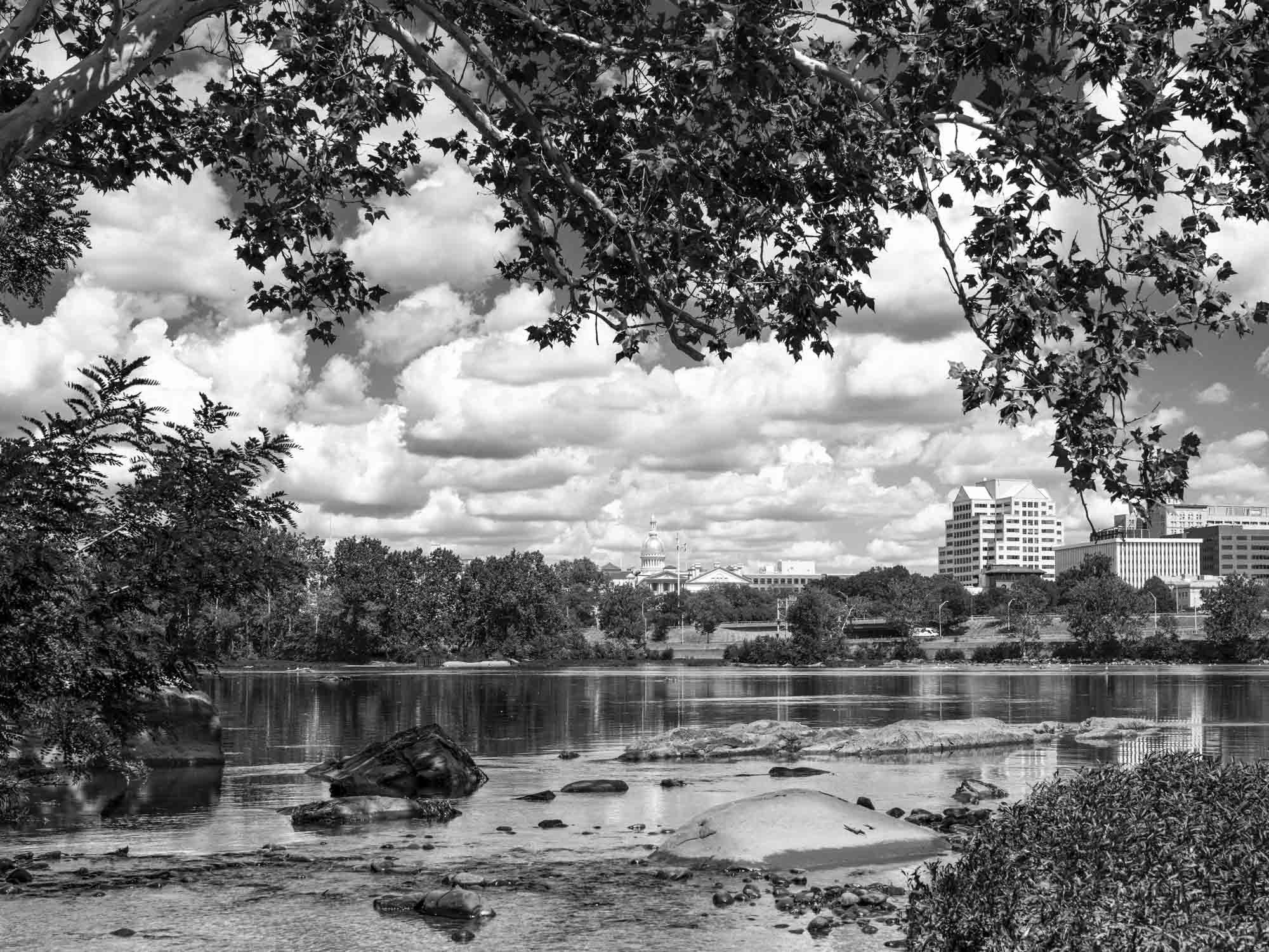 Black and white photo of a city skyline across a river with rocks and trees in the foreground and a cloudy sky.