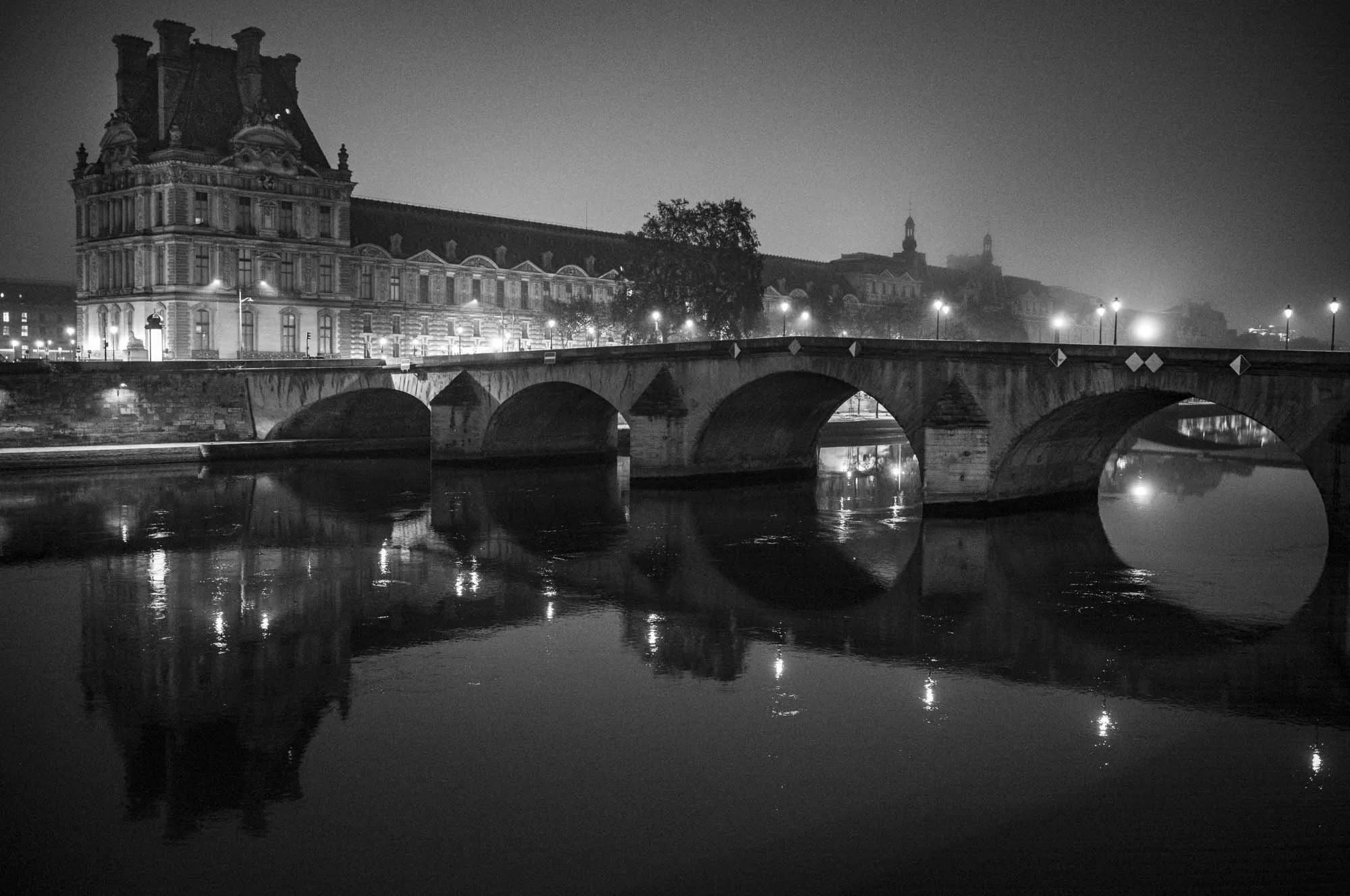 A nighttime black and white photograph of a historic building beside a bridge over a river, with streetlights illuminating the scene and reflections visible in the water.