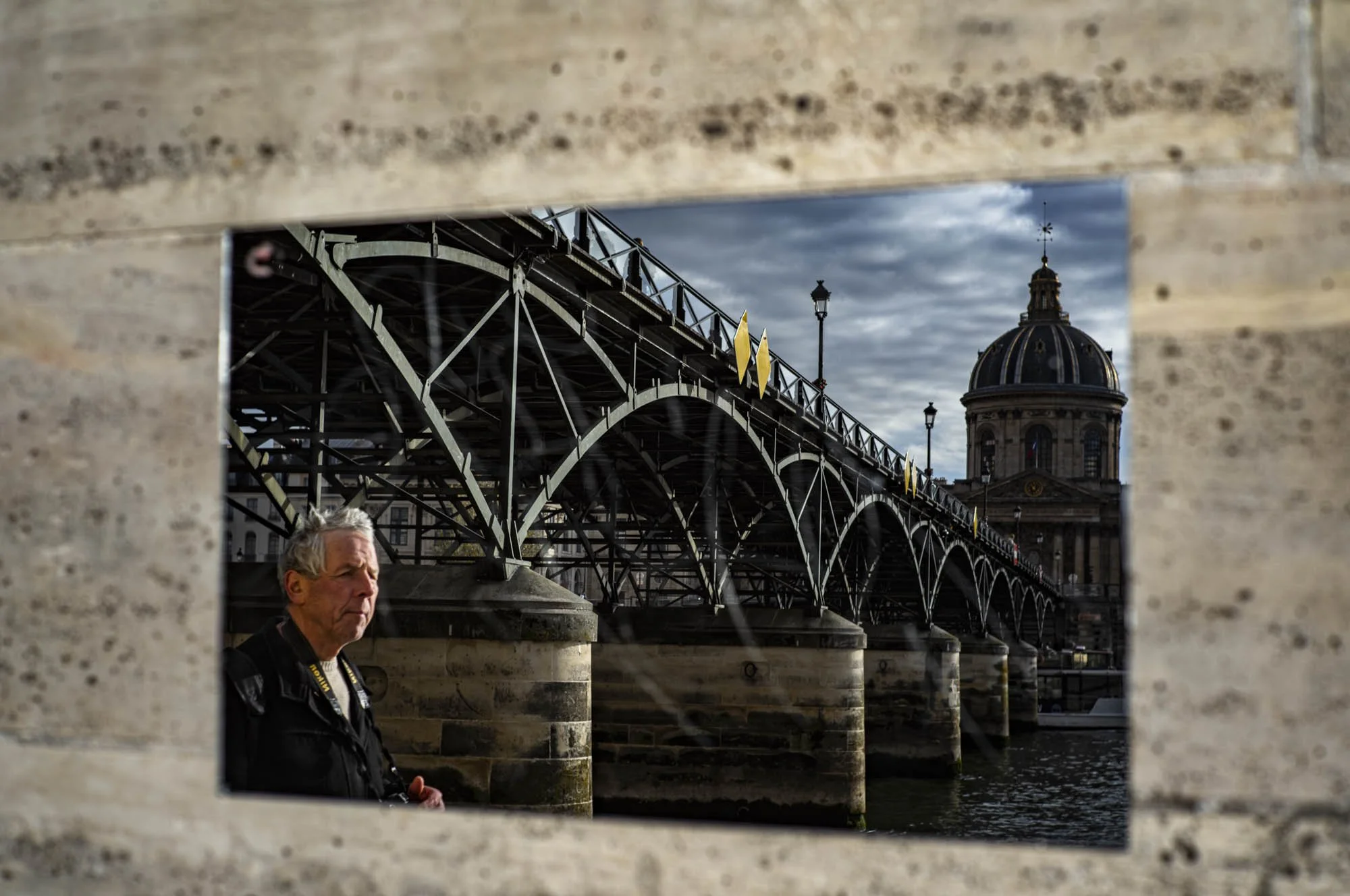 View of an old bridge over a river, with a historic building topped with a dome in the background, and a man sitting on the riverbank in the foreground, seen through a rectangular opening in a brick wall.