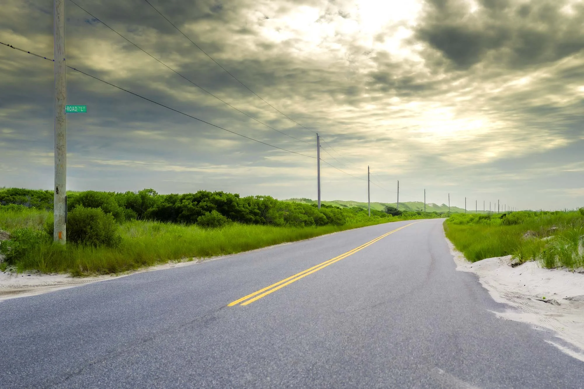 Empty rural road with yellow dividing lines, green grassy landscape, wooden utility poles along the side, and a cloudy sky.