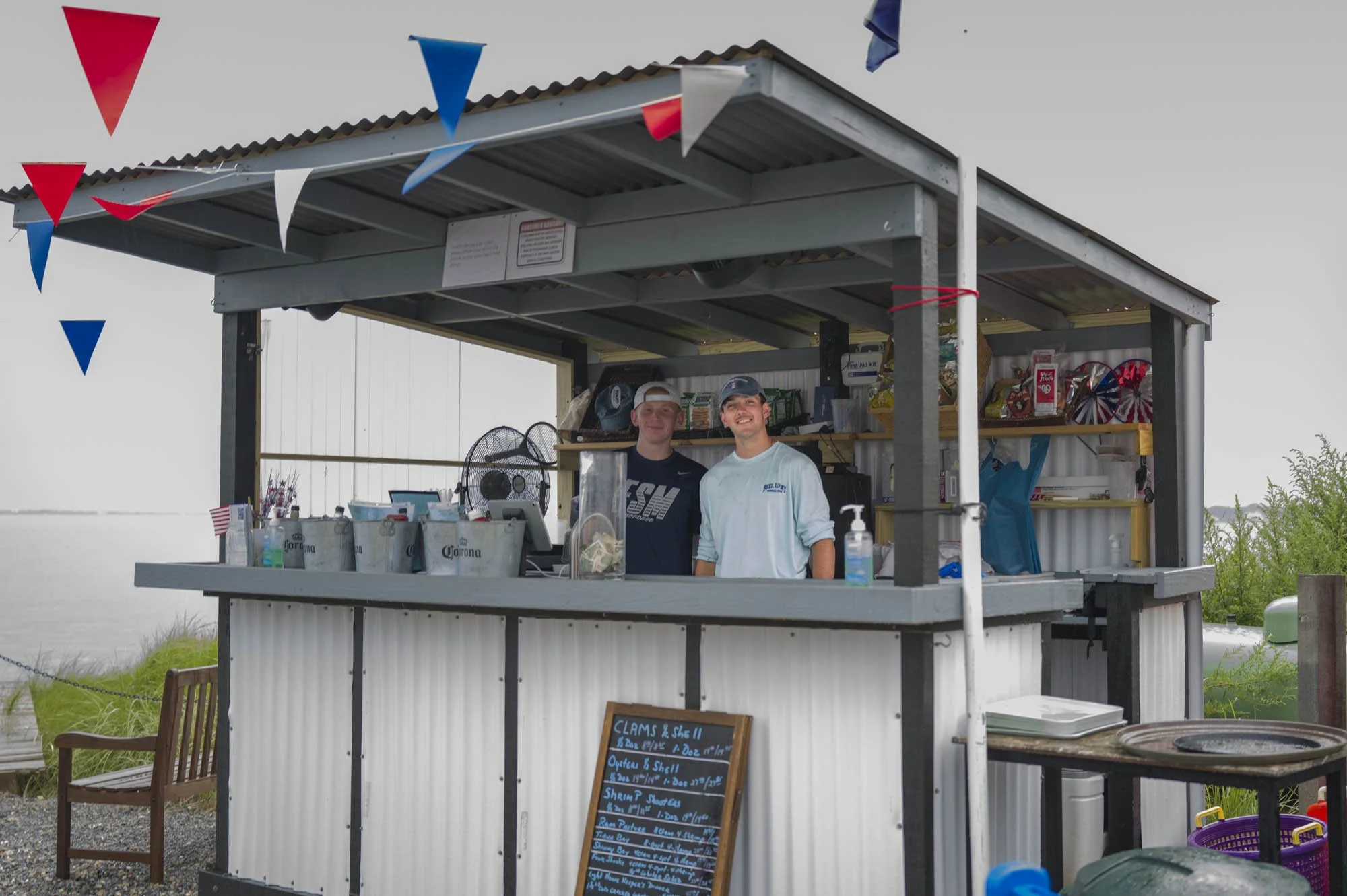 Two young men stand behind a portable seafood stand with a menu board, supplies, and decorations, near a body of water.