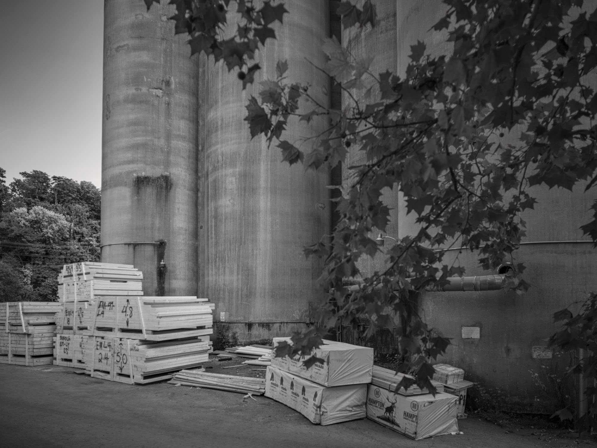 Construction supplies like lumber and insulation stored near a large industrial concrete silo, with tree branches and leaves partially covering the view.