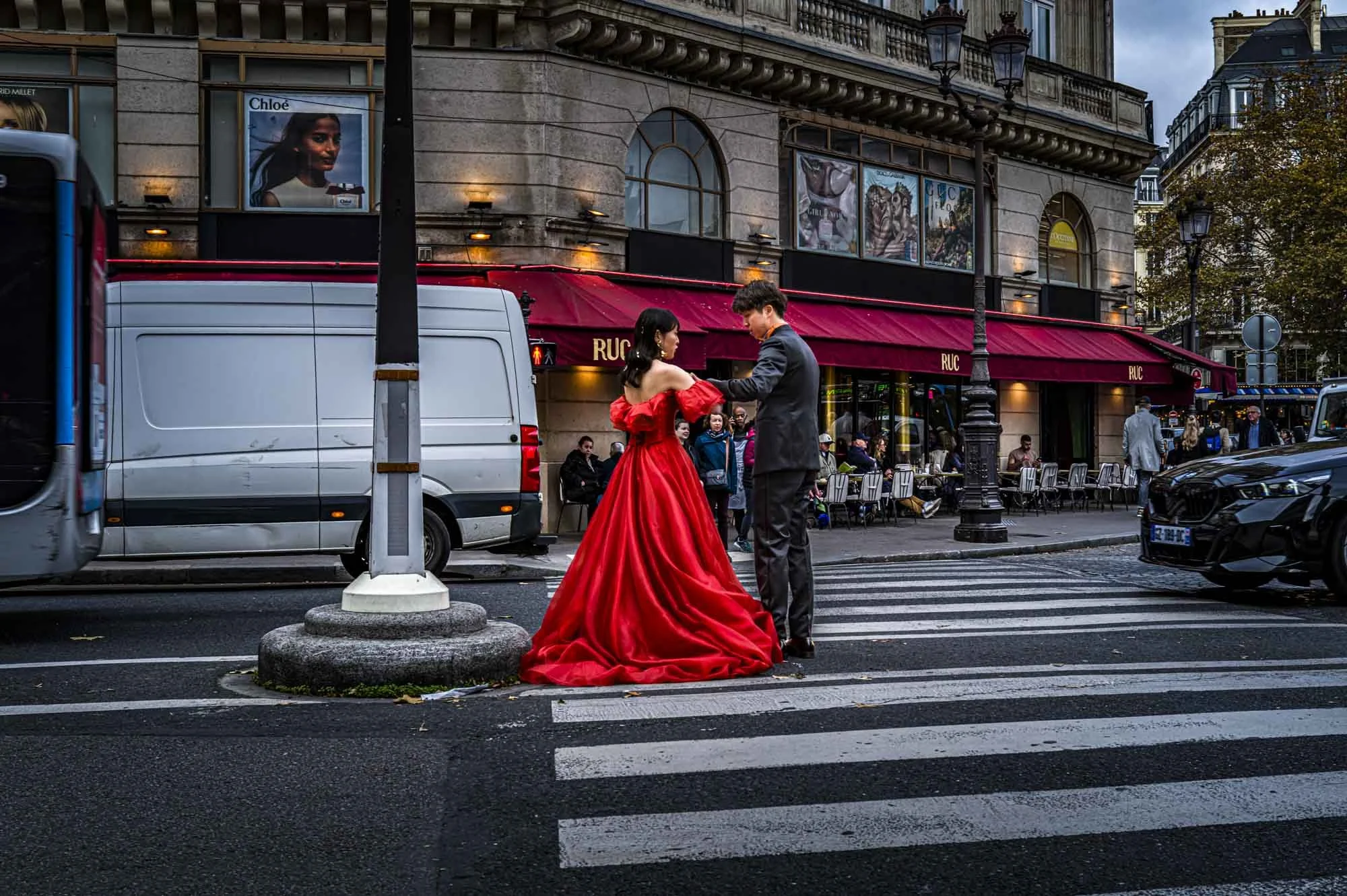 A couple dancing on a city street crosswalk, woman in a long red dress and man in a suit, with a café and pedestrians in the background.