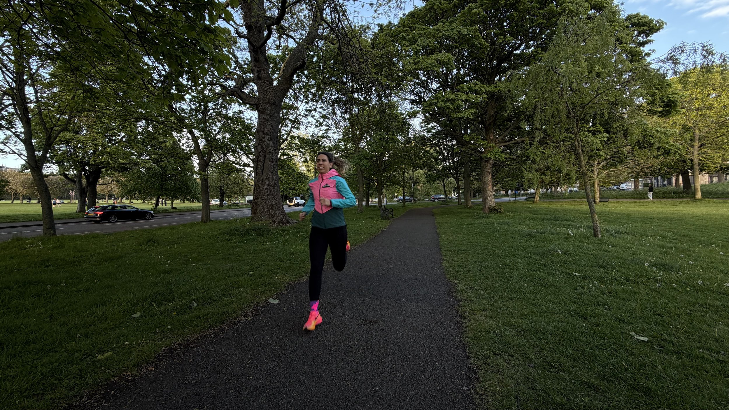 A woman jogging in a park surrounded by trees, wearing a pink and blue jacket, black pants, and pink running shoes.