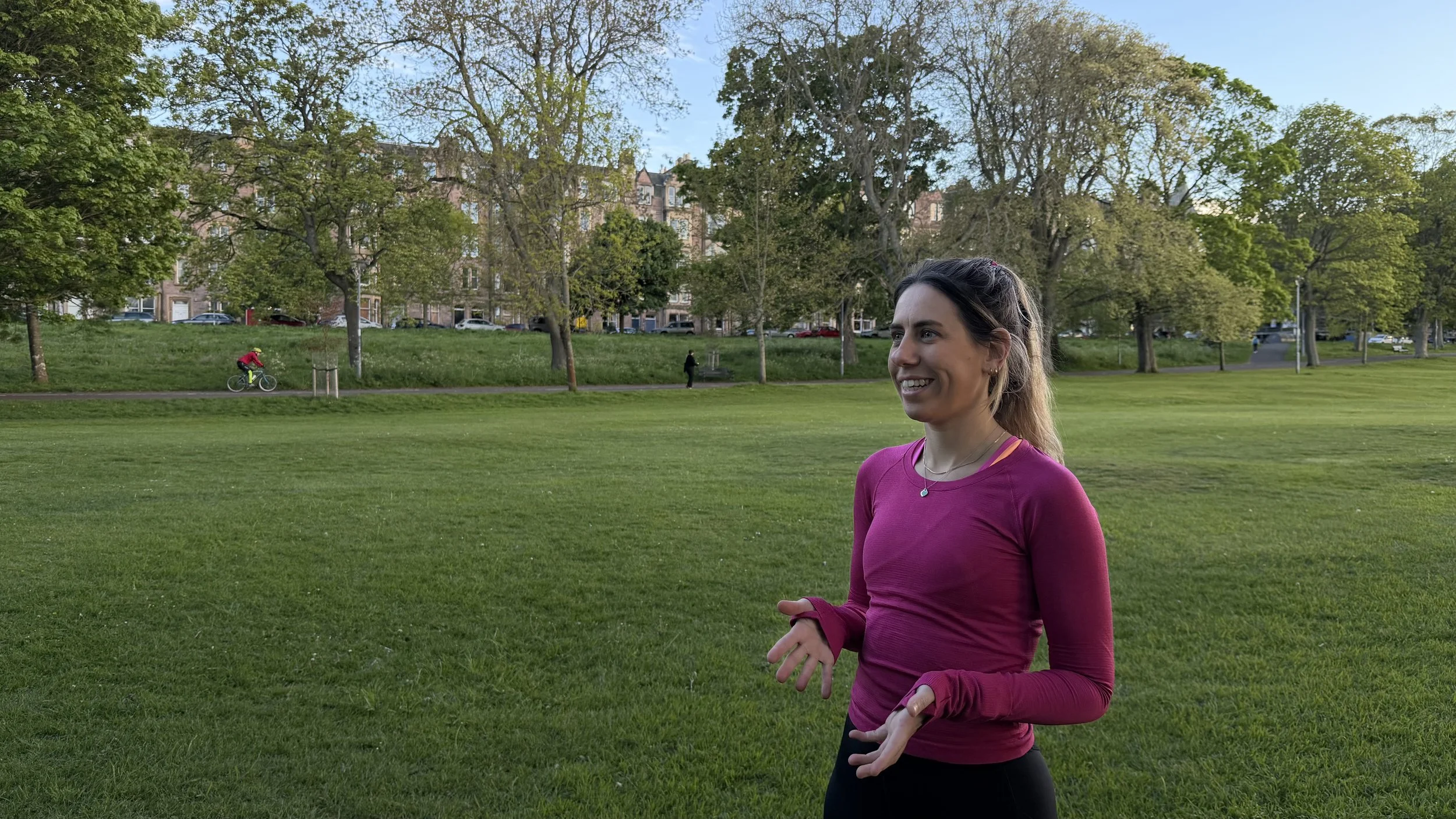 A woman jogging outdoors on a grassy field, smiling, wearing a pink long-sleeve shirt and black pants, with trees and buildings in the background.