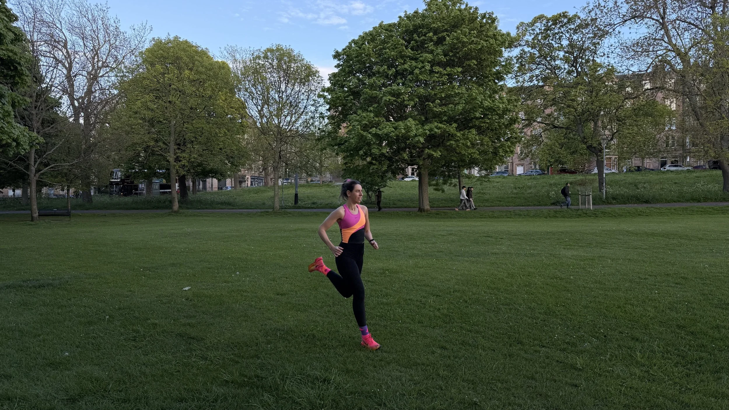 A woman in a pink sports bra and black leggings running in a park with green grass and trees, some with full foliage and some with no leaves, on a clear day with a blue sky.