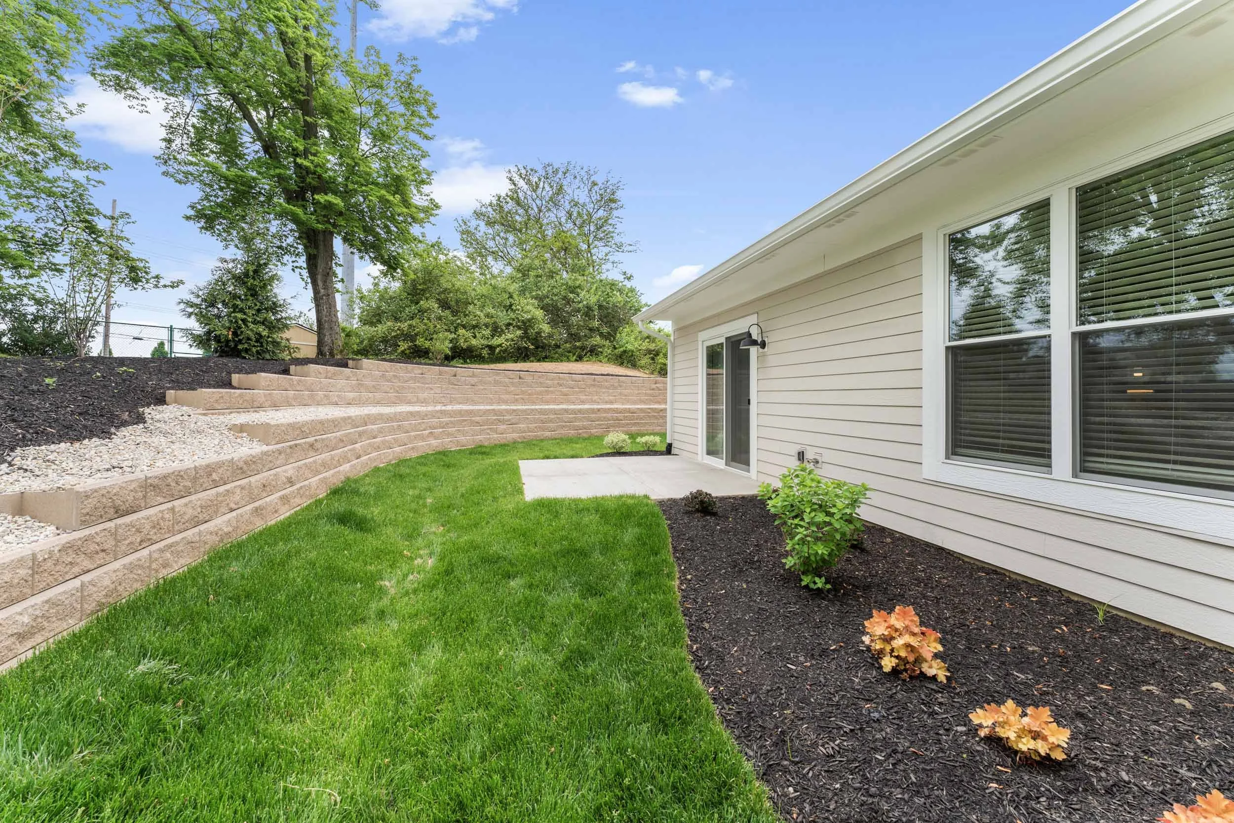 Backyard with a green lawn, landscaped flower bed with plants and mulch, beige house siding, a sliding glass door, and large windows. There are trees and a blue sky with a few clouds.