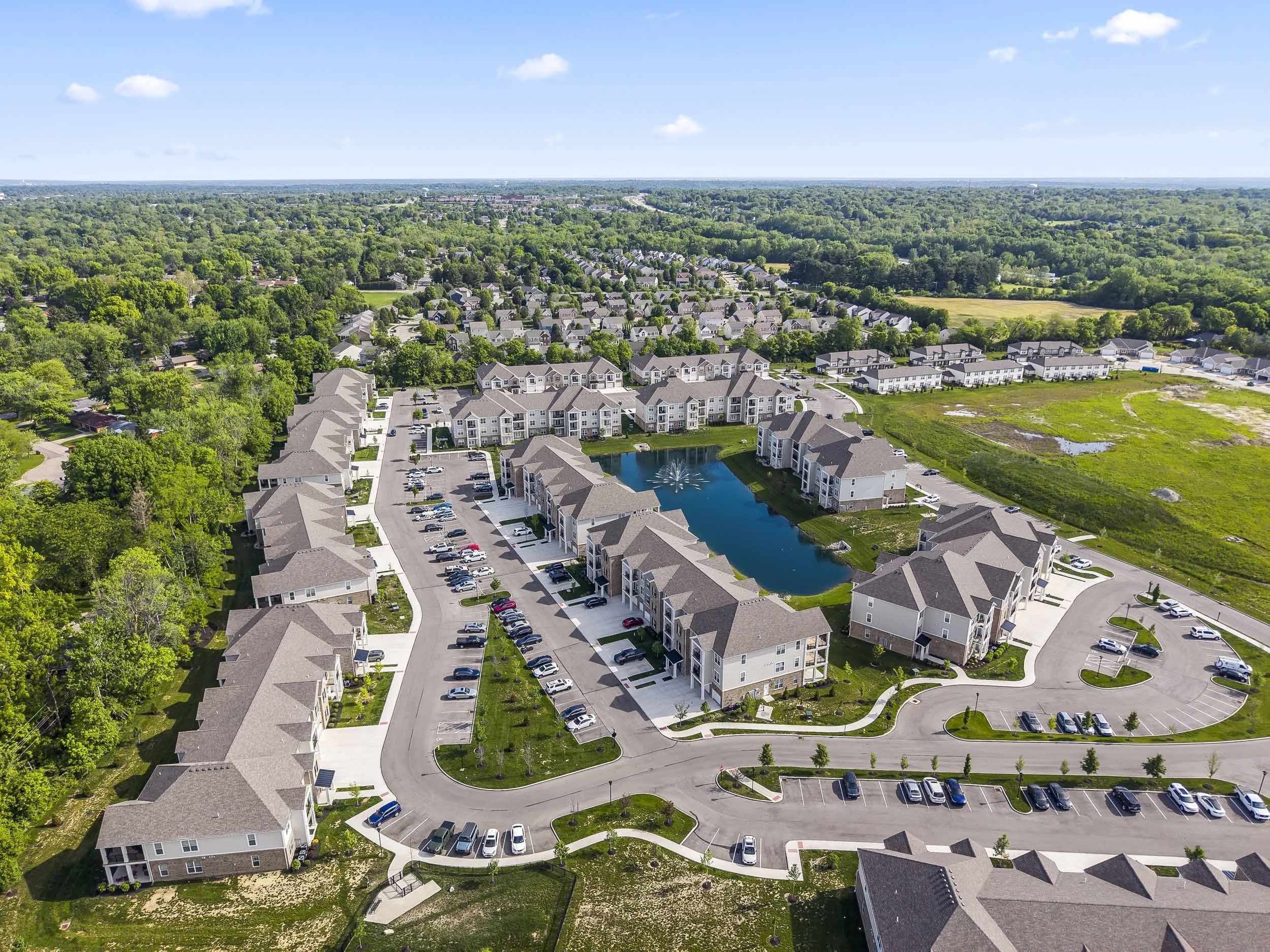 An aerial view of a residential neighborhood with multi-story apartments, a pond with a fountain, parking lots, and surrounded by trees and green fields under a blue sky with some clouds.