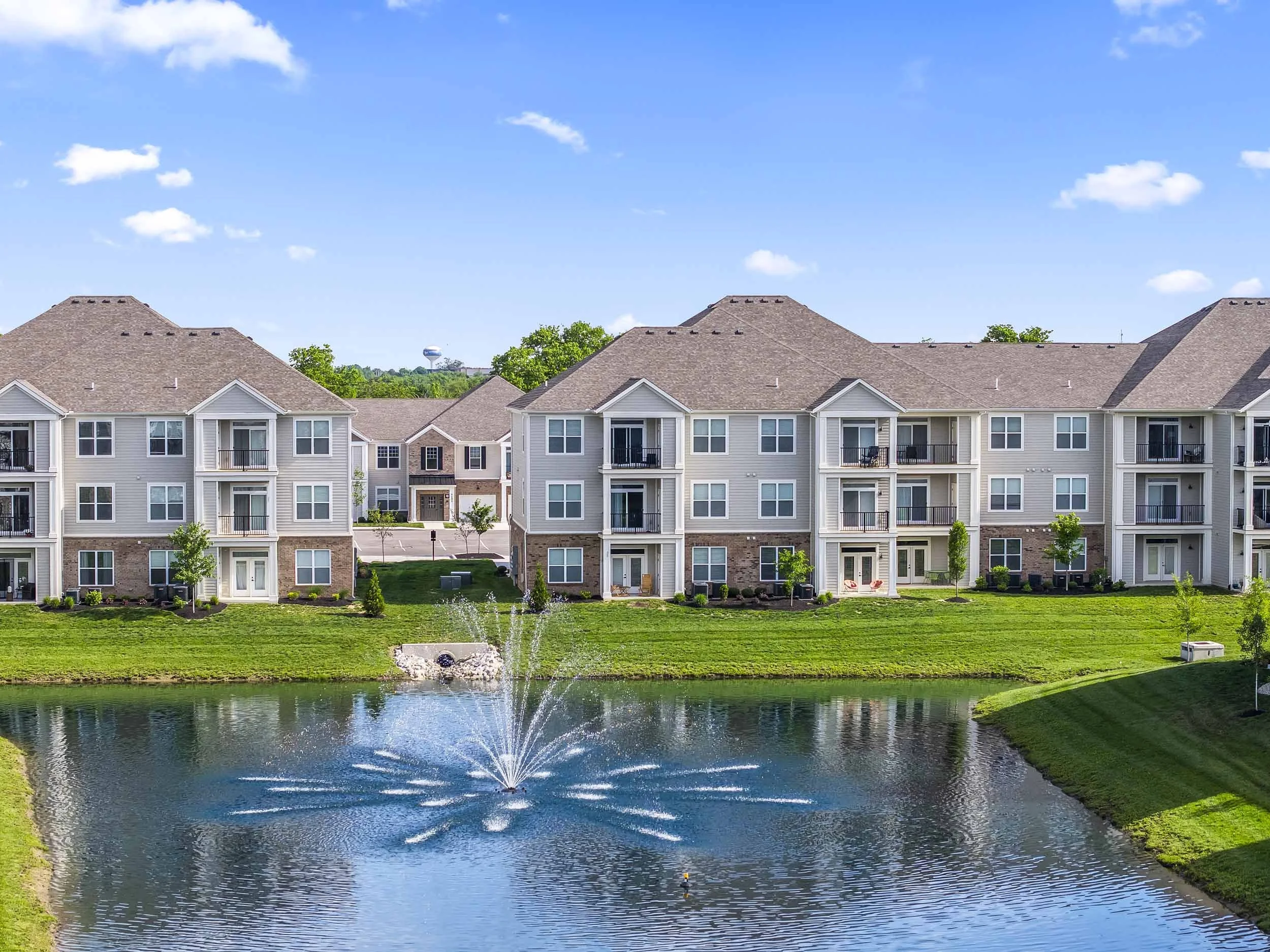A row of multistory residential buildings facing a pond with a fountain, under a blue sky with scattered clouds.