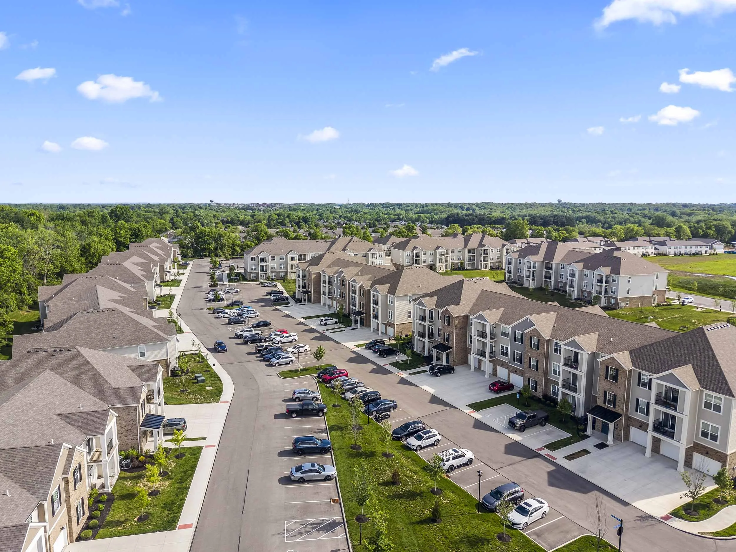 An aerial view of a residential apartment complex with multiple buildings, parking lots filled with cars, and surrounded by lush green trees, under a blue sky with a few scattered clouds.