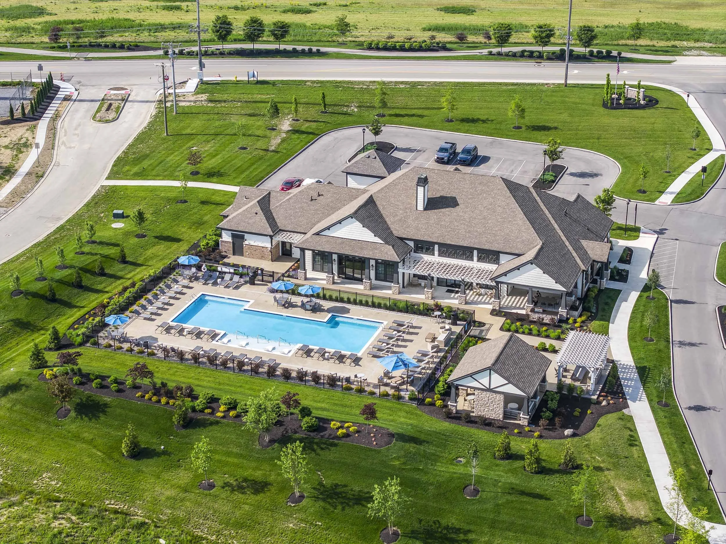 An aerial view of a residential area showing a house with a swimming pool, surrounded by well-maintained green grassy lawns, small trees, and paved walkways. There is a parking lot with several parked cars and a circular driveway in front of the hous