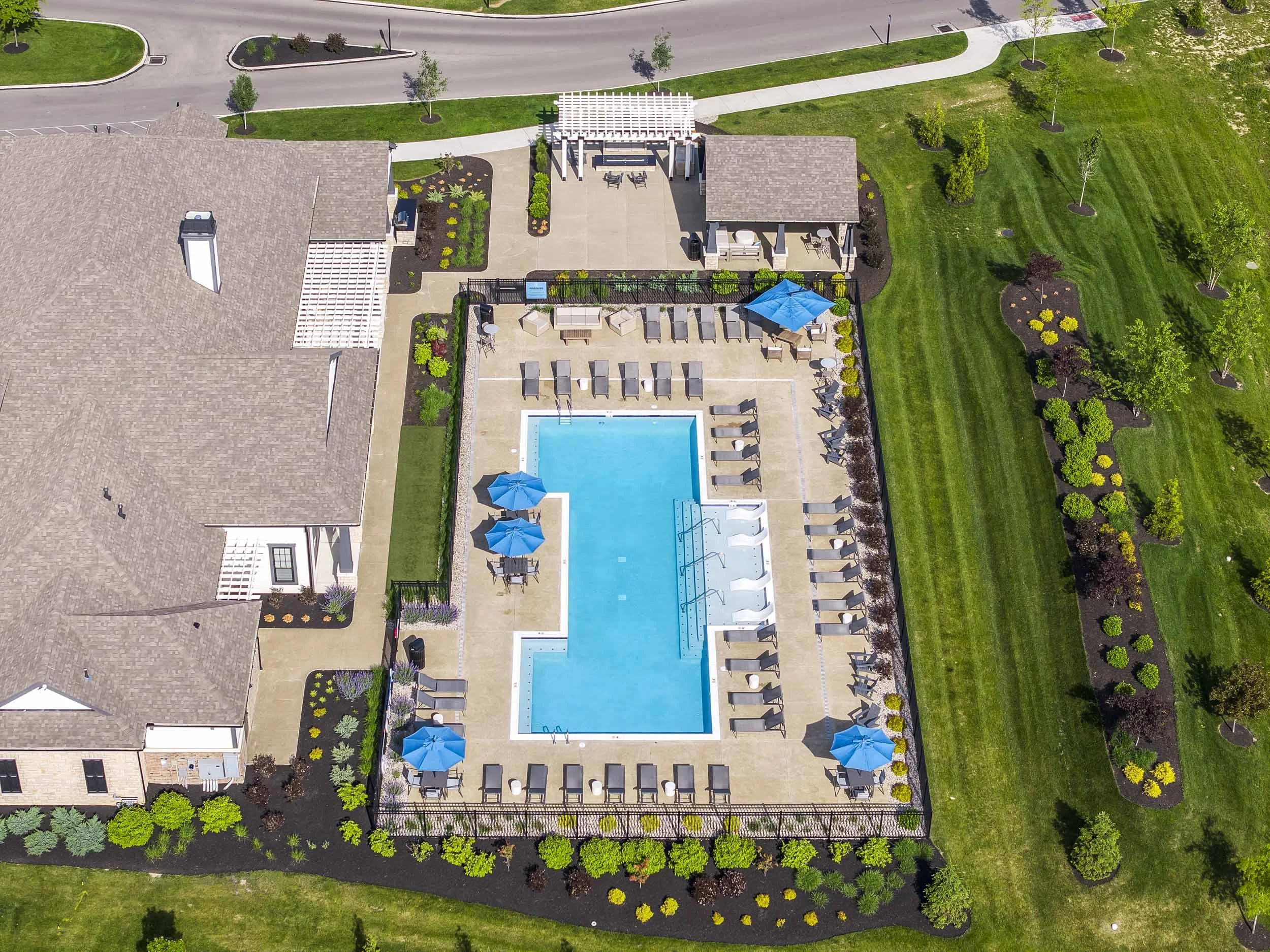 Aerial view of a swimming pool area with lounge chairs and umbrellas, adjacent to a house with a large porch, surrounded by landscaped gardens and green grass.