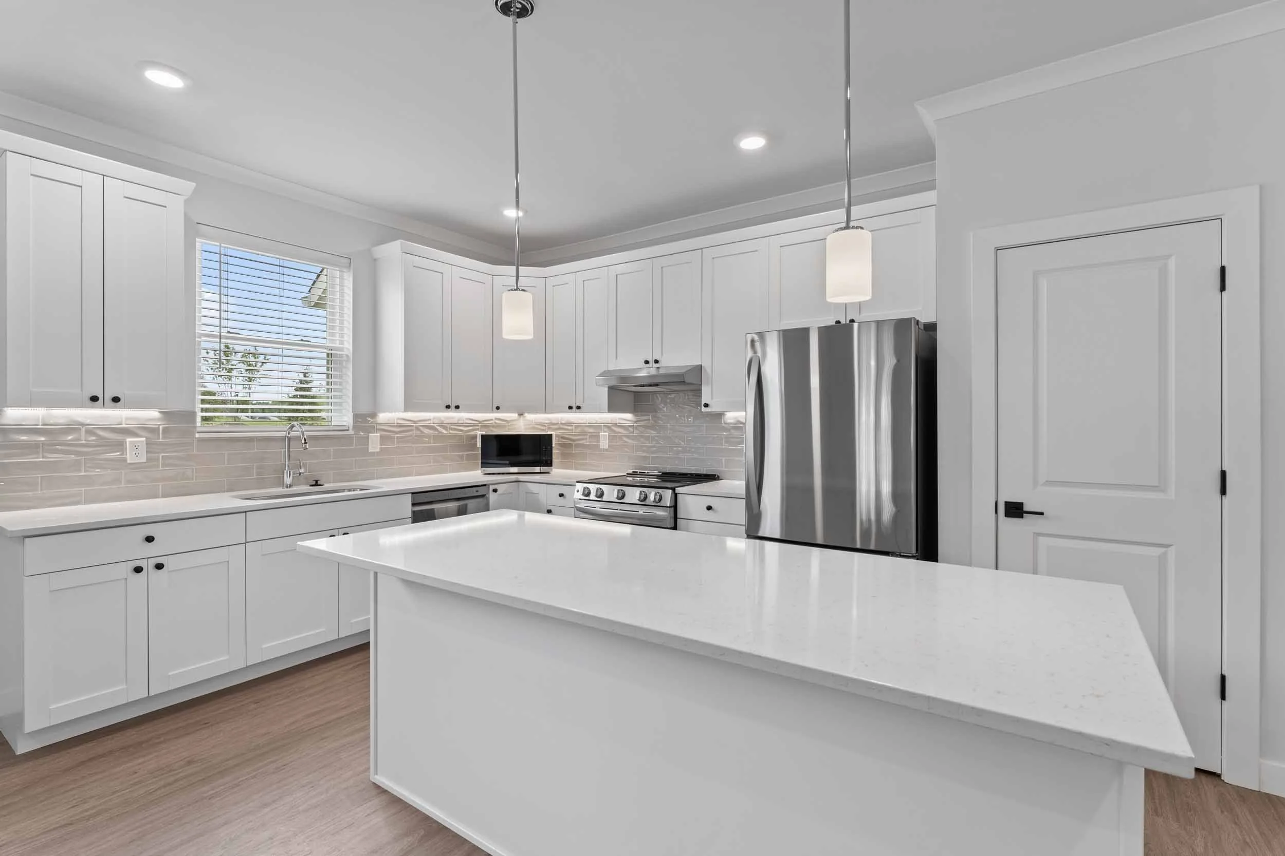 Modern white kitchen with ample cabinet space, stainless steel appliances, a small microwave, and a window with blinds letting in natural light. Two pendant lights hang above the island, and there is a gray tile backsplash.
