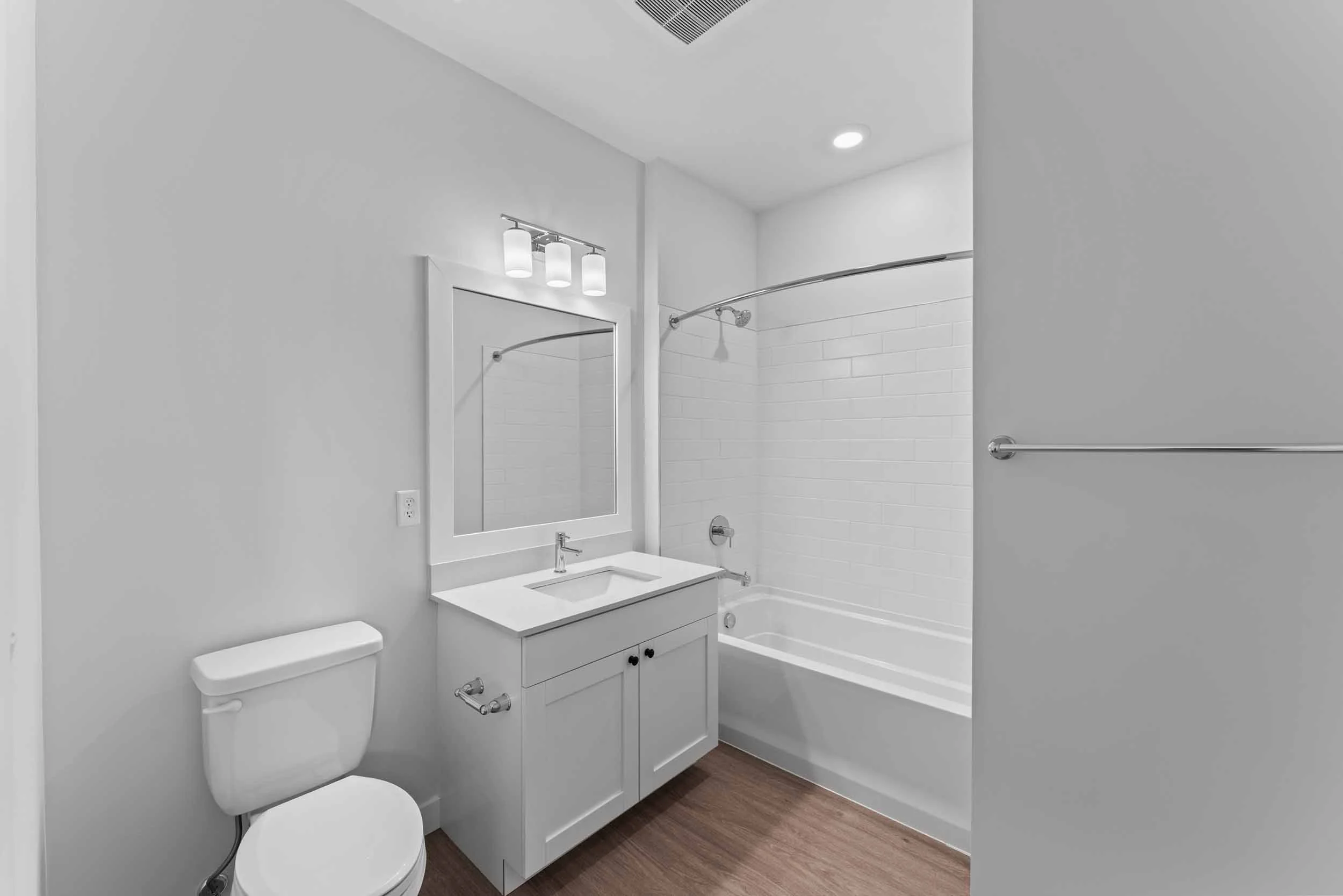 A modern, minimalist bathroom featuring a white toilet, a white vanity with a sink, a rectangular mirror, illuminated by a three-light fixture, with a bathtub and shower with a curved shower rod and white subway tile. The flooring is brown wood, and 