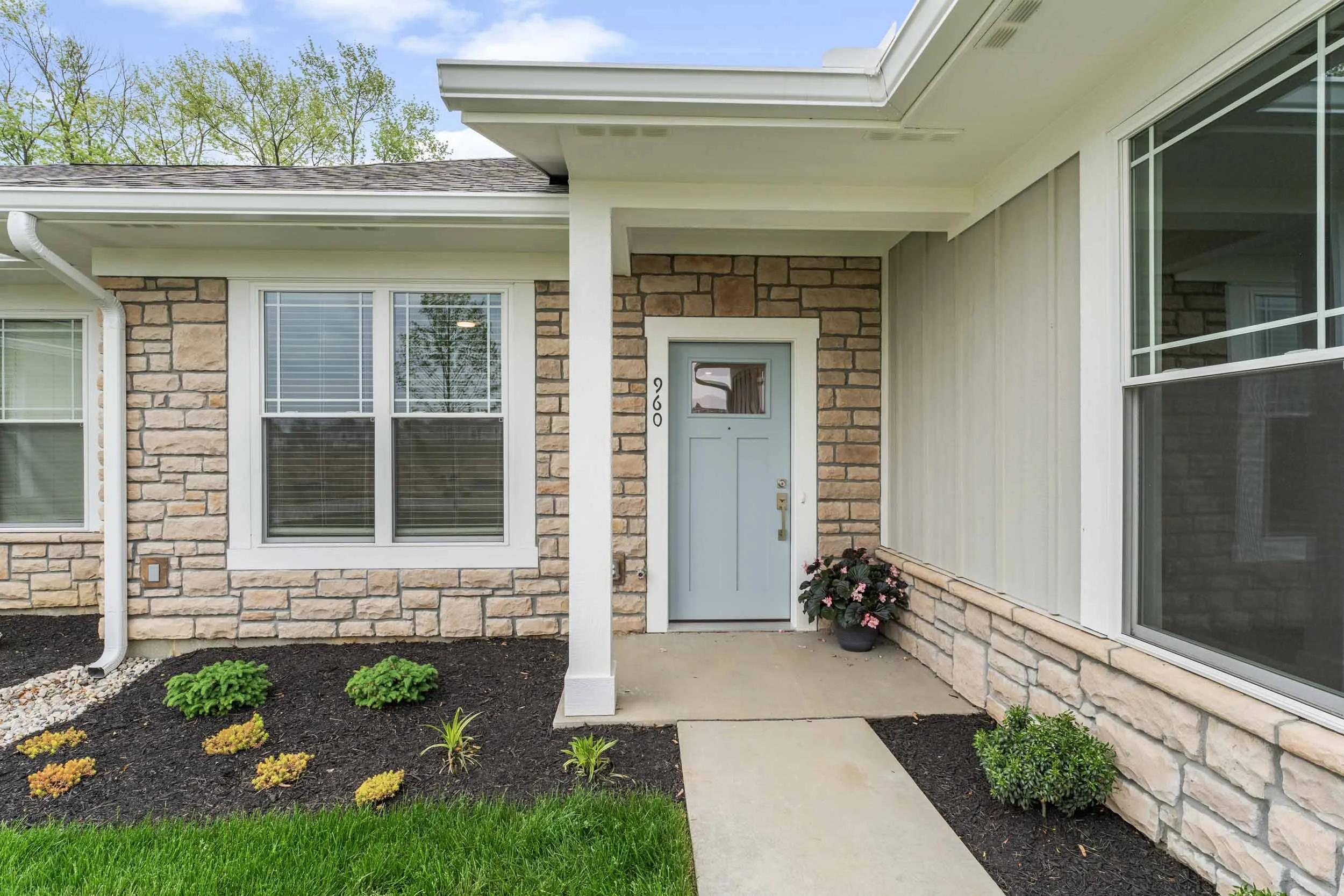 Front porch of a house with a light blue door, brick and siding exterior, windows with blinds, potted flowers, small plants, and a concrete walkway leading to the door.