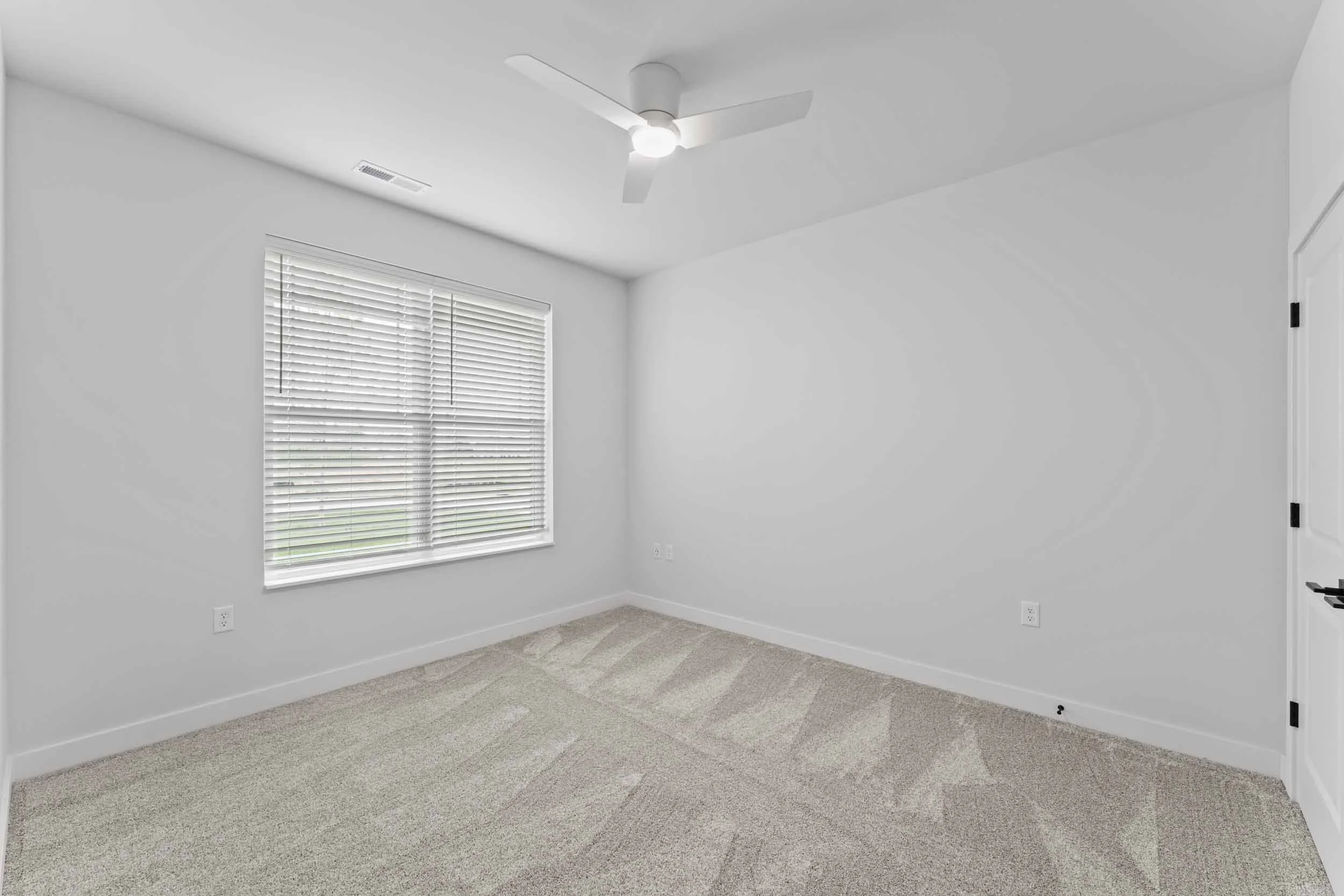 Empty room with a window covered by white blinds, ceiling fan with light, beige carpet, white doors, and white walls.