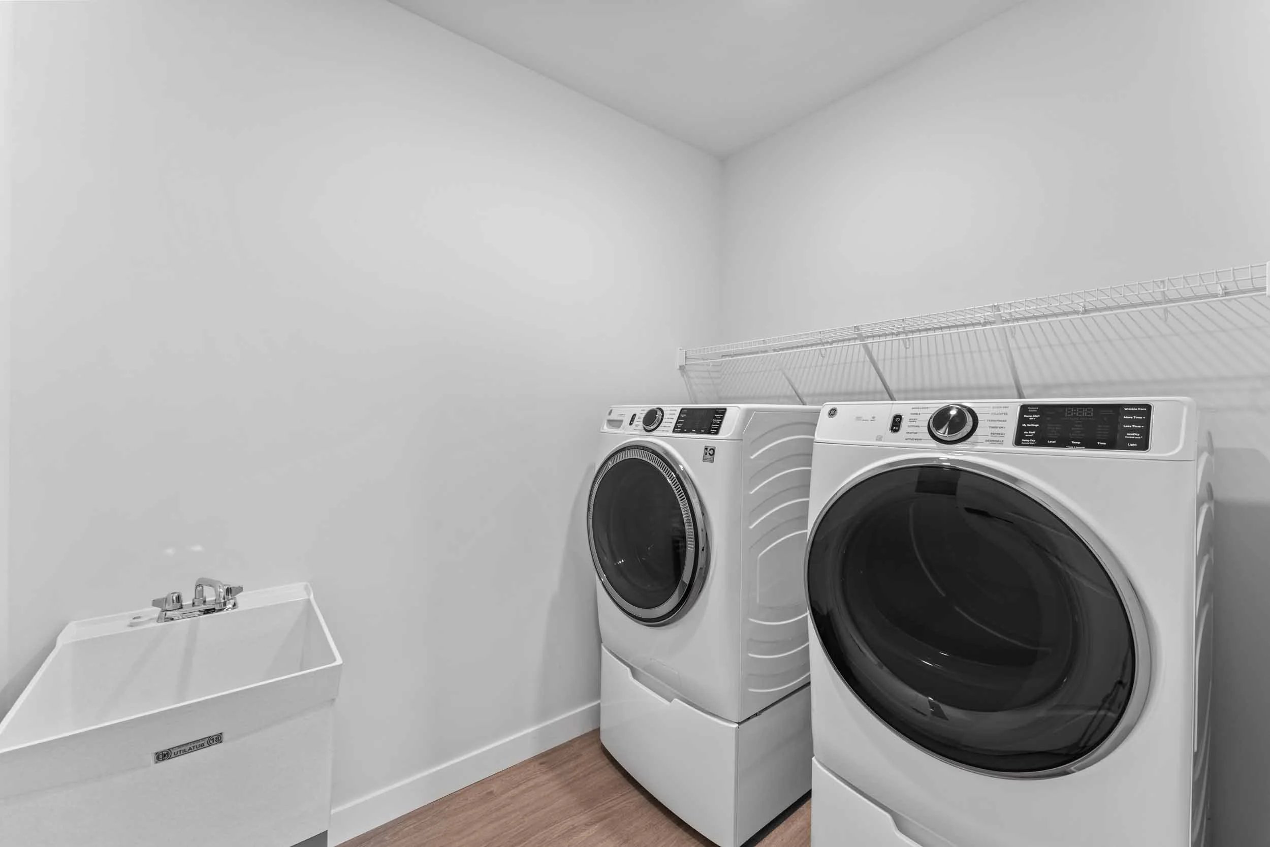 A laundry room with a washing machine and dryer side by side under a wire shelf, with a laundry sink on the left and hardwood flooring.