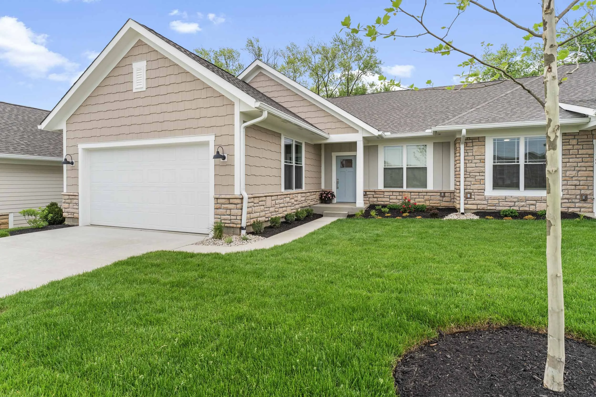 A modern house with a beige and stone exterior, a white garage door, and a small front porch surrounded by green grass and trees.