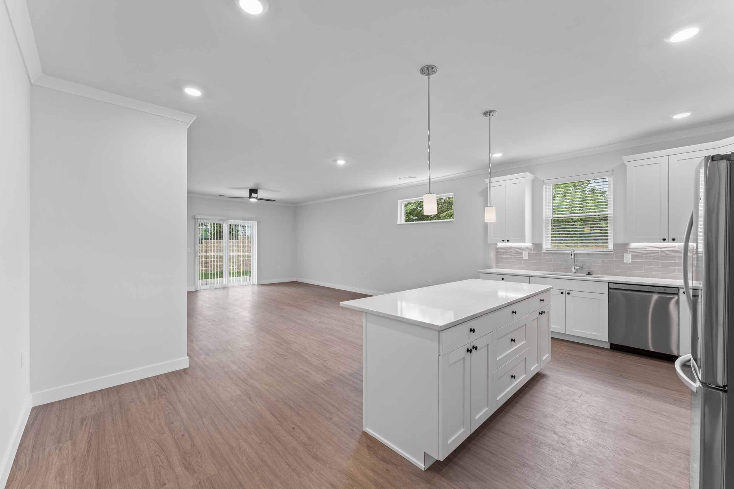 Empty modern kitchen with white cabinets, a marble island, stainless steel appliances, and large windows with blinds, overlooking a backyard.