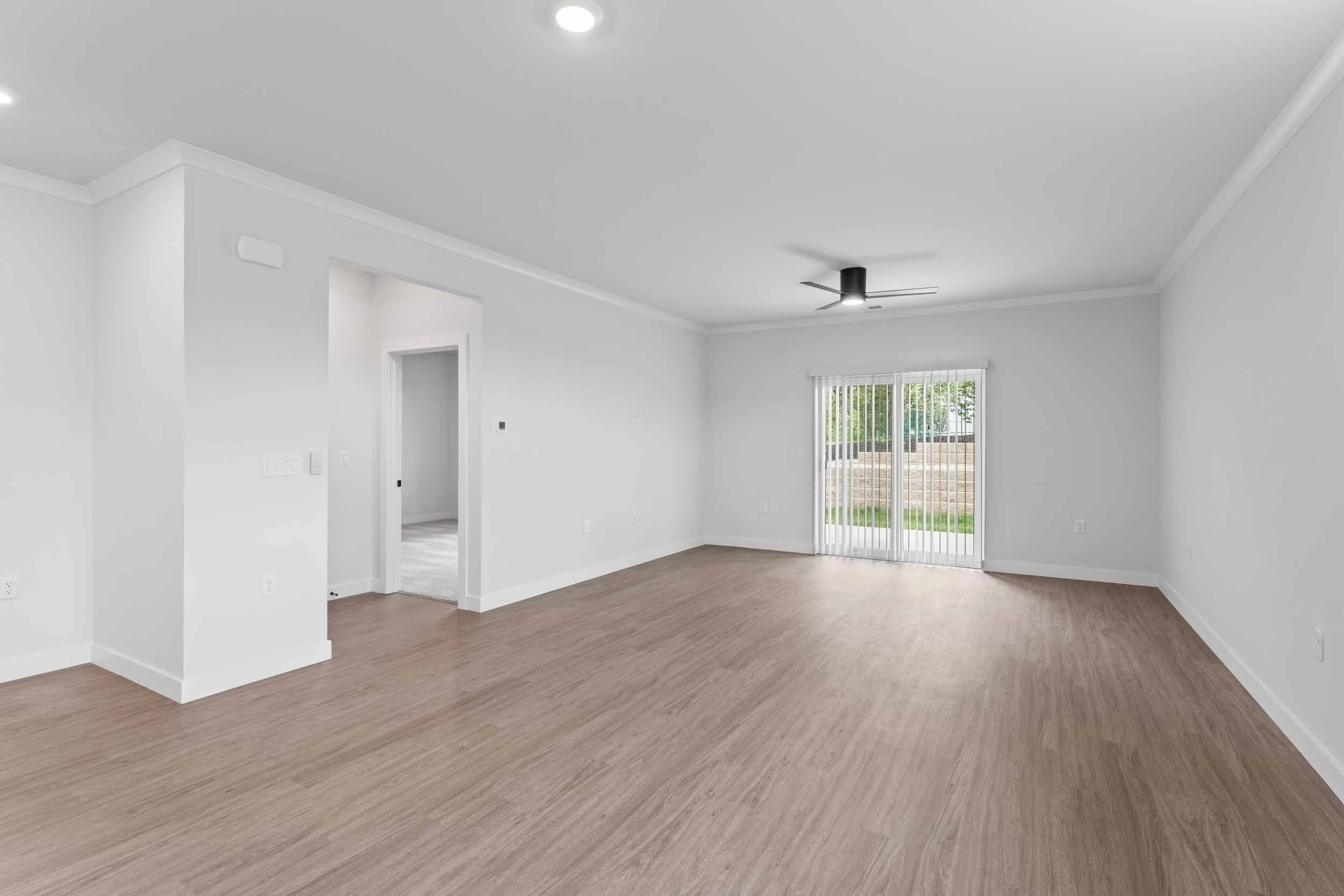 Empty living room with white walls, wood flooring, sliding glass door with vertical blinds, ceiling fan, and a small hallway leading to another room.