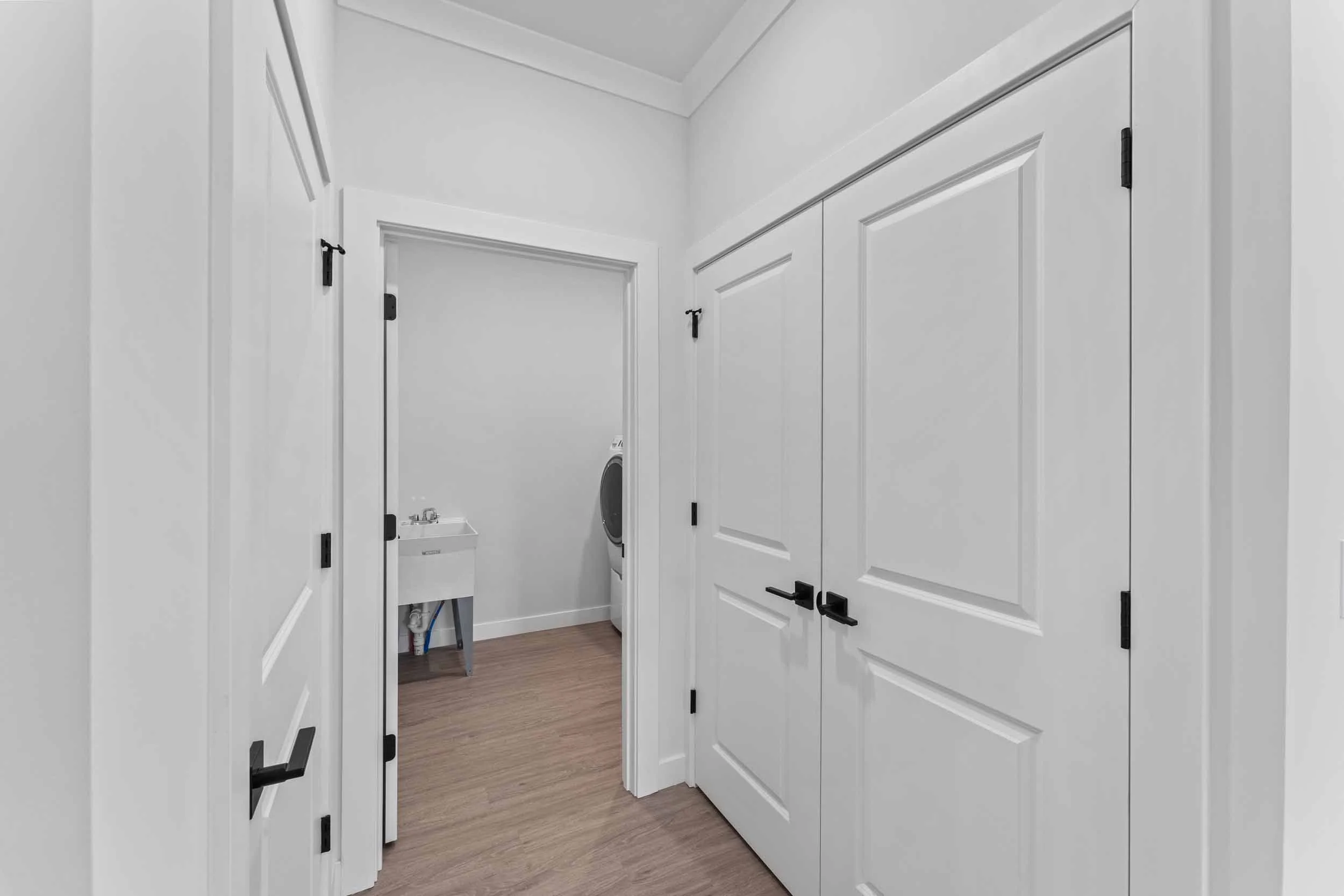 A laundry room with white walls, white cabinets, and a washer-dryer in the back corner. The floor is wood, and there is a small utility sink visible near the appliances.