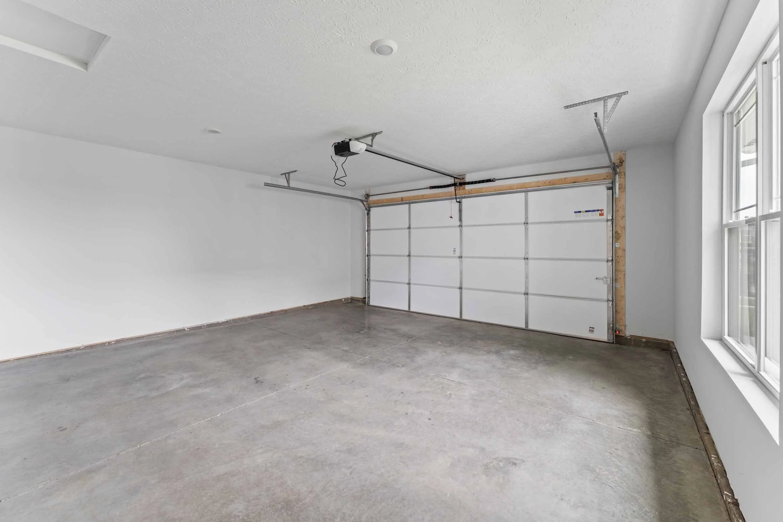 Empty garage with concrete floor, white walls, large window on the right, and a garage door with an automatic opener system on the ceiling.