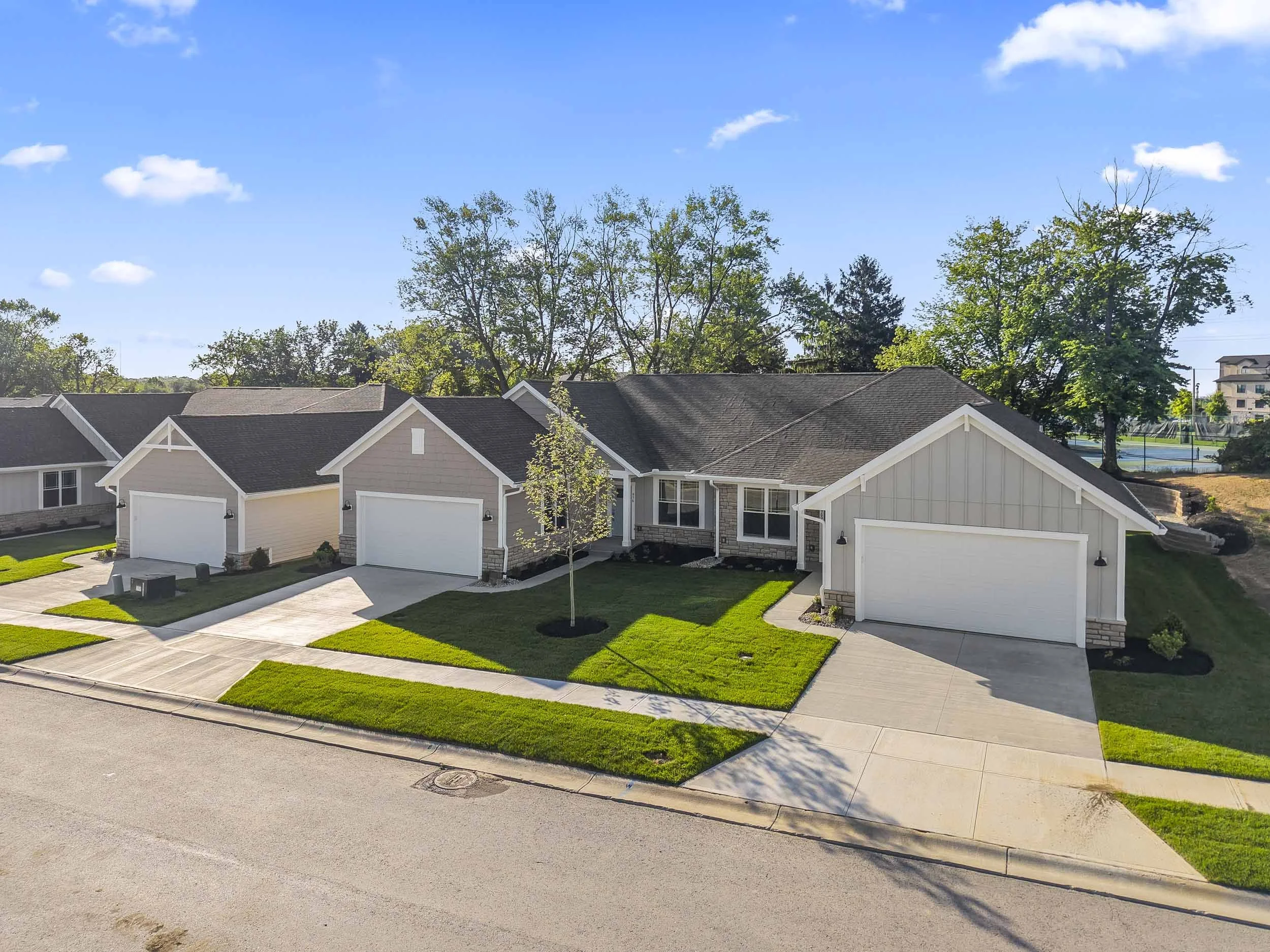 A suburban neighborhood with a row of modern houses featuring white and beige exteriors, dark shingled roofs, and manicured lawns. There are trees with green foliage and a clear blue sky with a few scattered clouds.