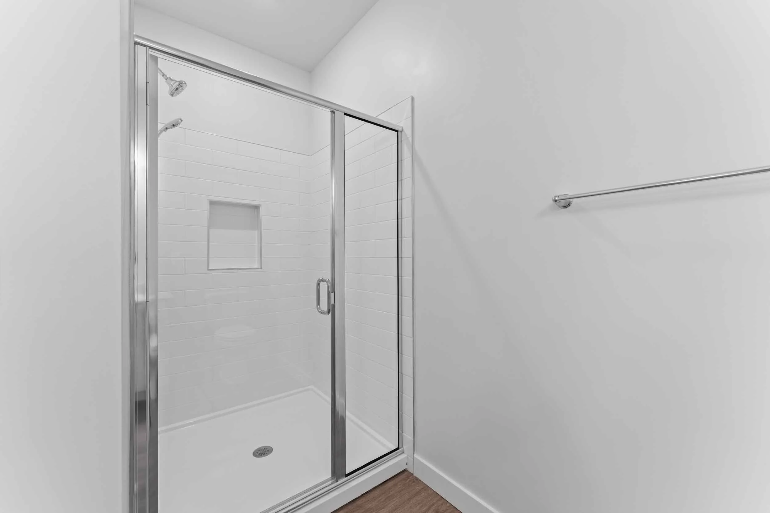 A modern walk-in shower with clear glass doors, white tiled walls, a built-in shower niche, and a rain showerhead, in a minimalist bathroom with gray walls and wood flooring.