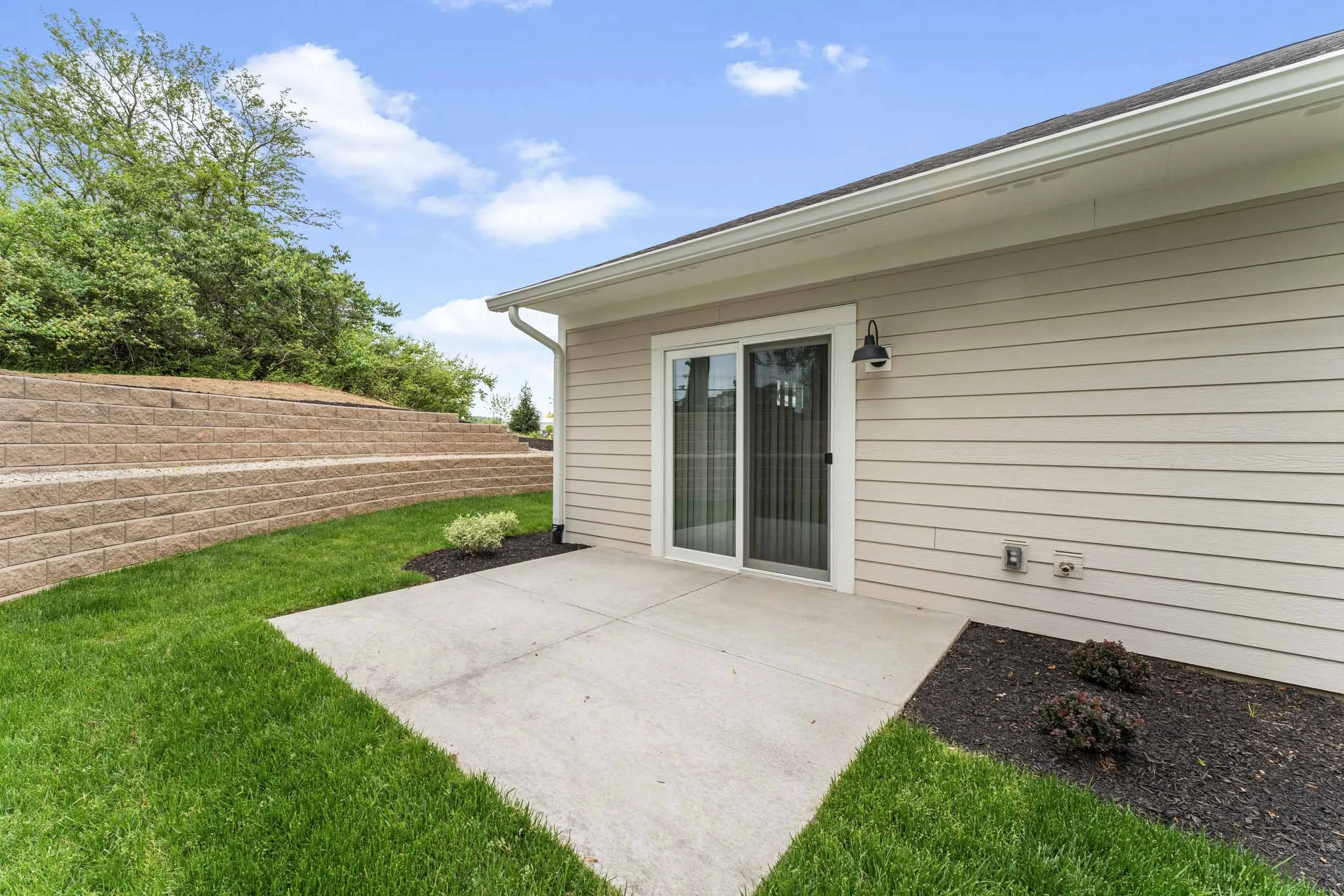 Backyard patio area with a sliding glass door, outdoor wall light, a small shrub, a well-maintained lawn, and a brick retaining wall bordering a grassy hill under a partly cloudy sky.