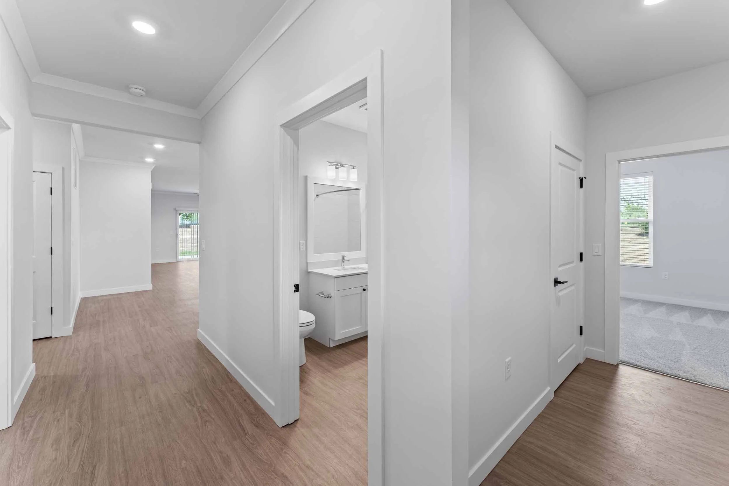 Interior of a modern house with white walls, wood flooring, a small bathroom with a sink and toilet, and multiple rooms visible through doorways and windows.