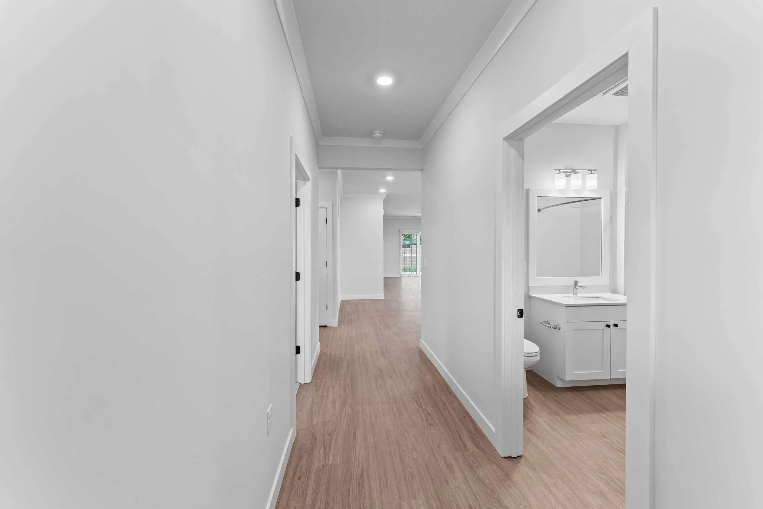empty hallway in a modern house, with white walls, wood flooring, and a bathroom with a mirror and vanity on the right side