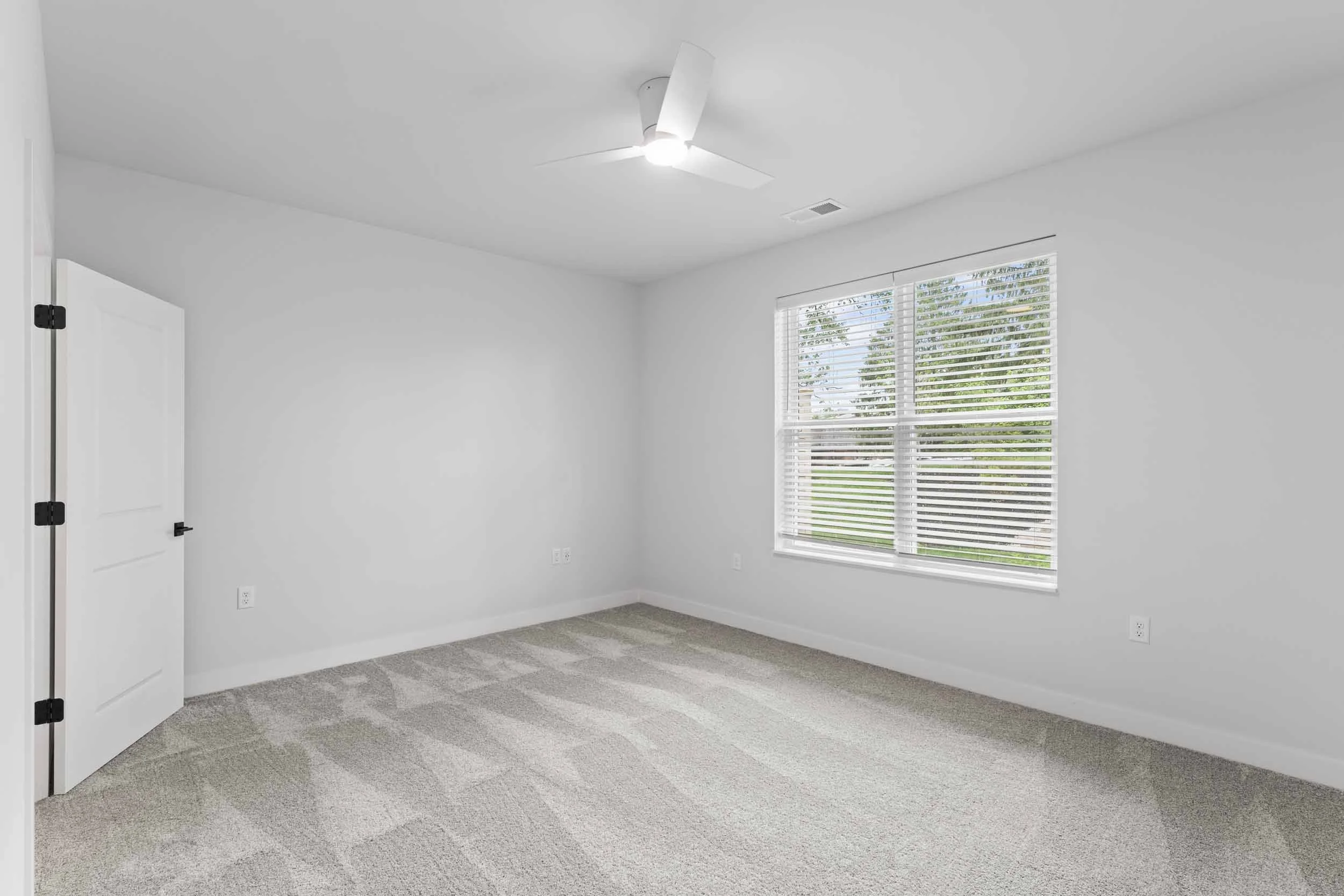 A bright empty bedroom with white walls, a white ceiling fan, a window with white blinds, light gray carpet, and white baseboards.