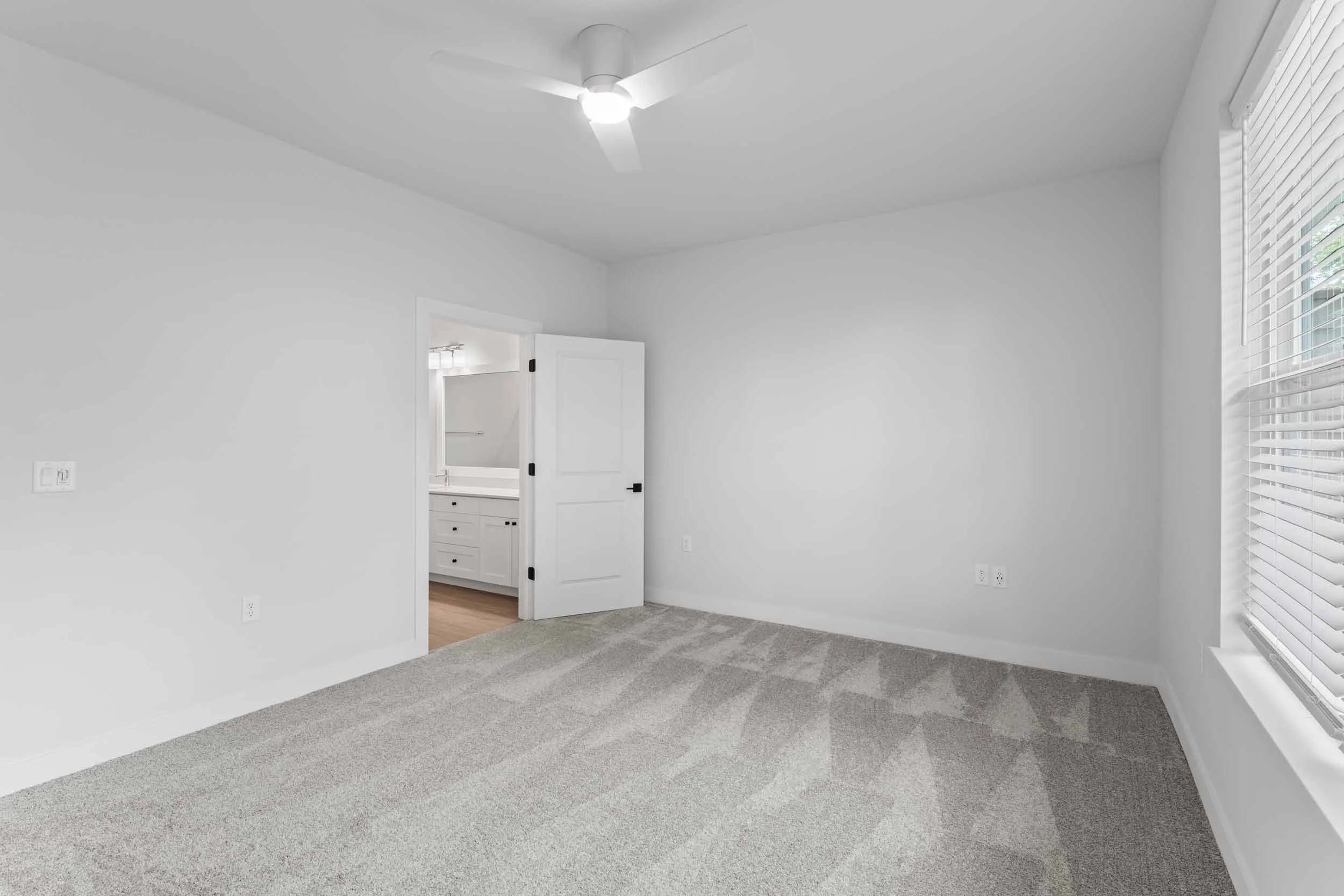 Empty room with gray carpet, white walls, a ceiling fan, a large window with blinds, and an open door leading to a bathroom with a white vanity and sink.