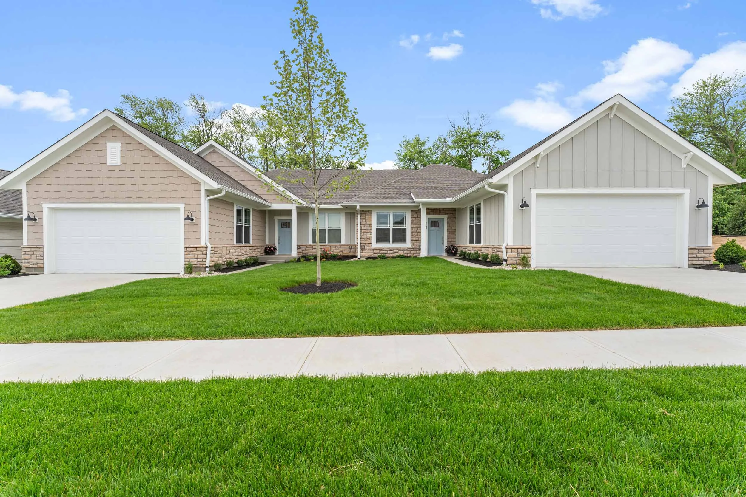 Front view of a modern suburban house with two attached garages, a small tree in the front yard, and a well-maintained green lawn under a clear blue sky.
