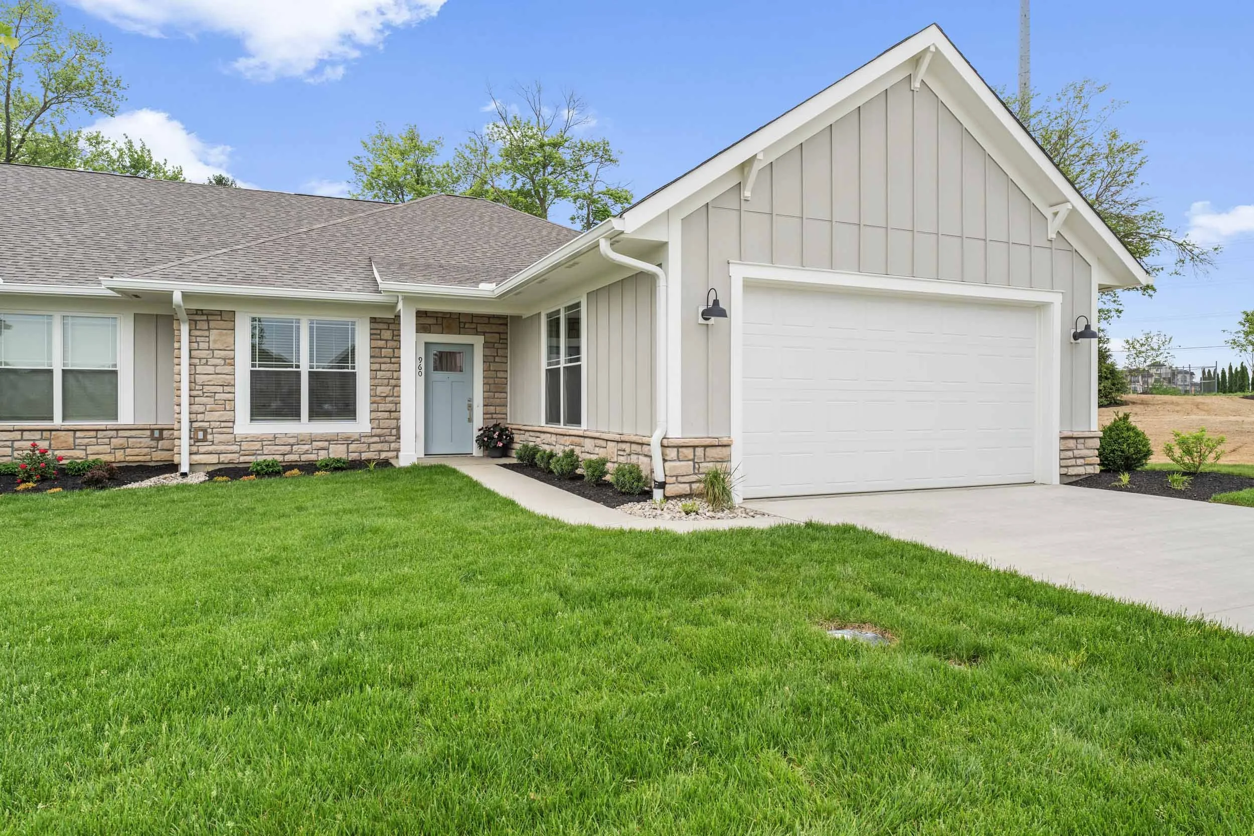 A suburban house with a well-maintained green lawn, brick and siding exterior, a single door, and a large attached garage door, set under a partly cloudy blue sky.